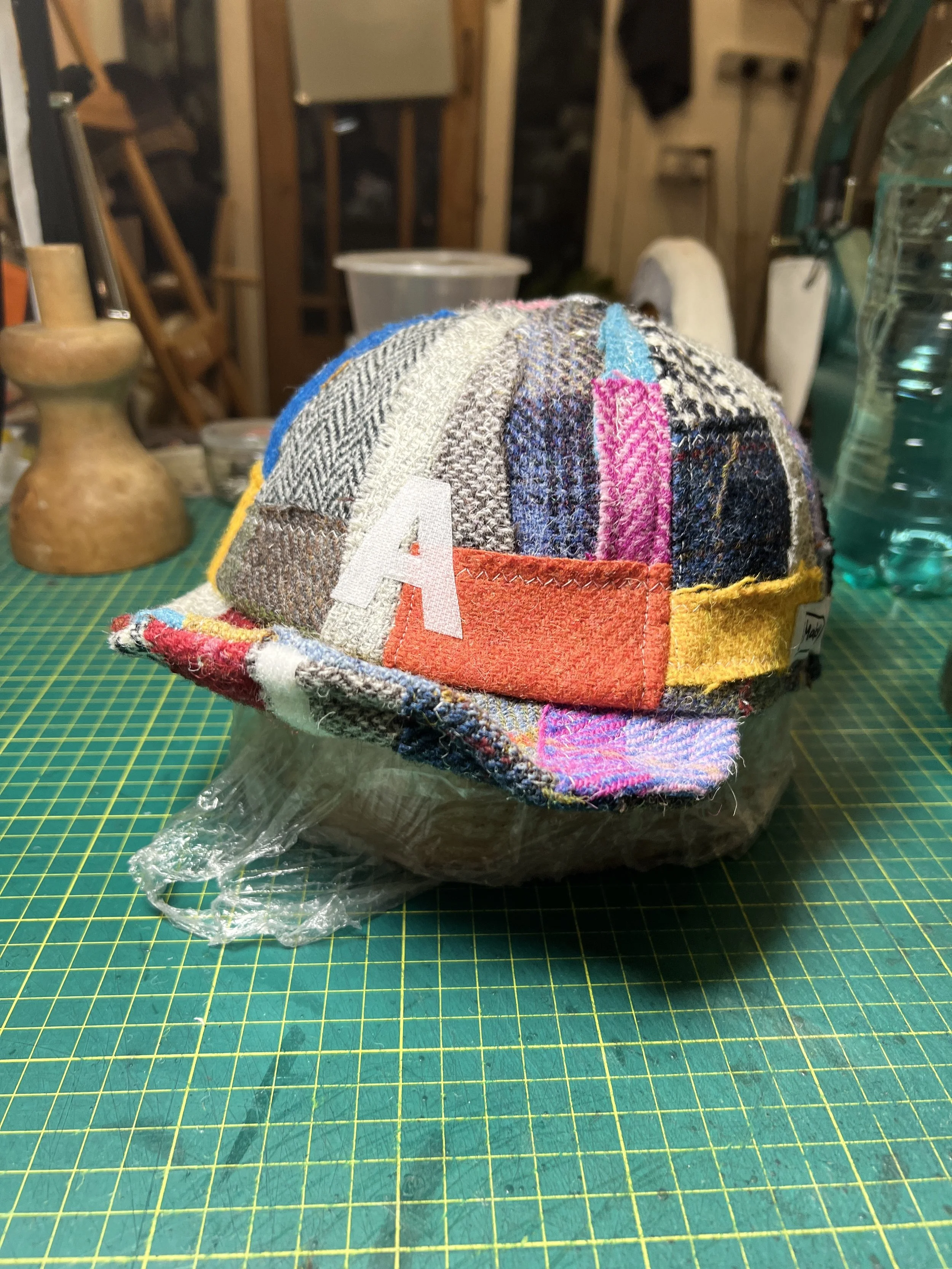 A one-of-a-kind Makshy patchwork cap, personalized with the letter 'A', displayed in the heart of the design studio. The vibrant, multi-colored tweed hat is set against a backdrop of a milliner's tools and work in progress.