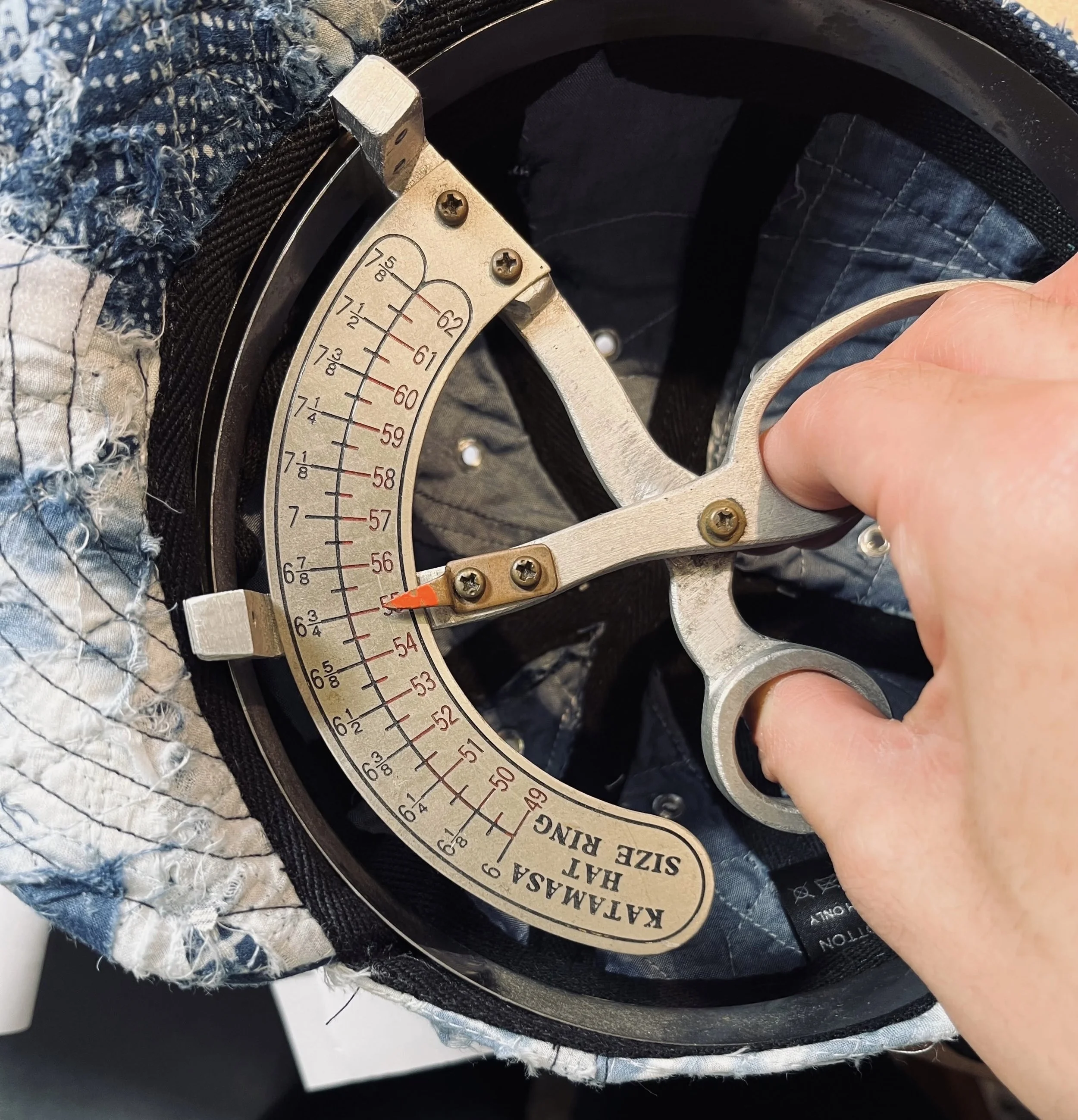 The precision craftsmanship of Makshy Millinery. A milliner uses a vintage-style hat sizing tool to measure the interior of a quilted, Boro-style patchwork cap, ensuring a perfect fit.