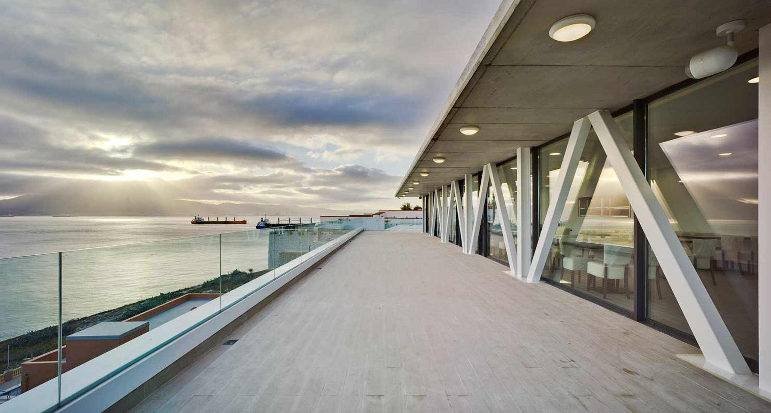 Modern building terrace with glass railing overlooking the ocean at sunset, with two ships in the distance and cloudy sky.