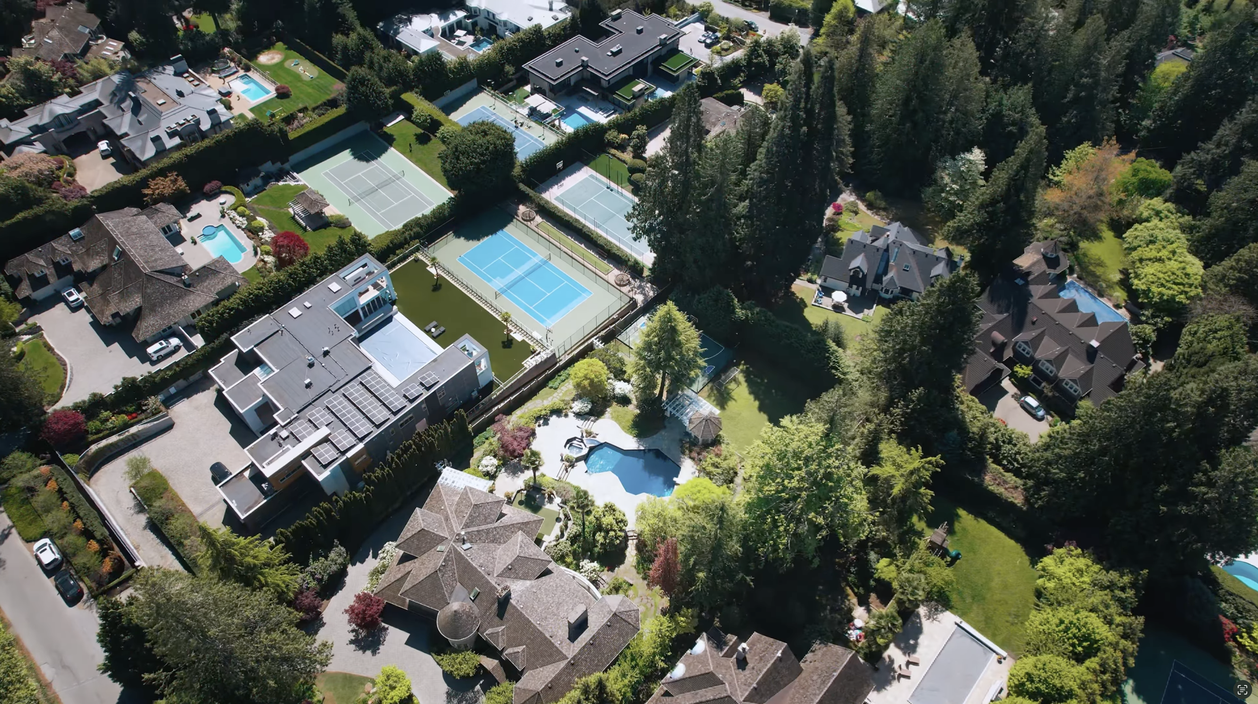 Aerial view of a residential neighborhood with multiple houses, some with pools, tennis courts, and green yards, surrounded by trees.