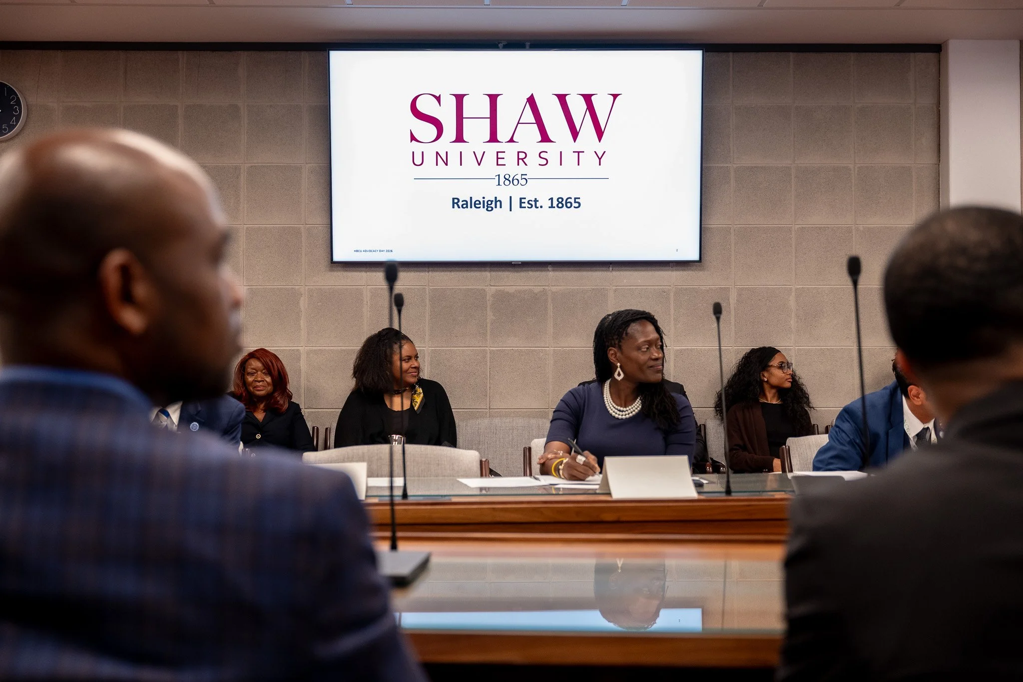 A group of diverse people sitting at a conference table during a meeting at Shaw University, with a large screen displaying the university's name and founding date in the background.