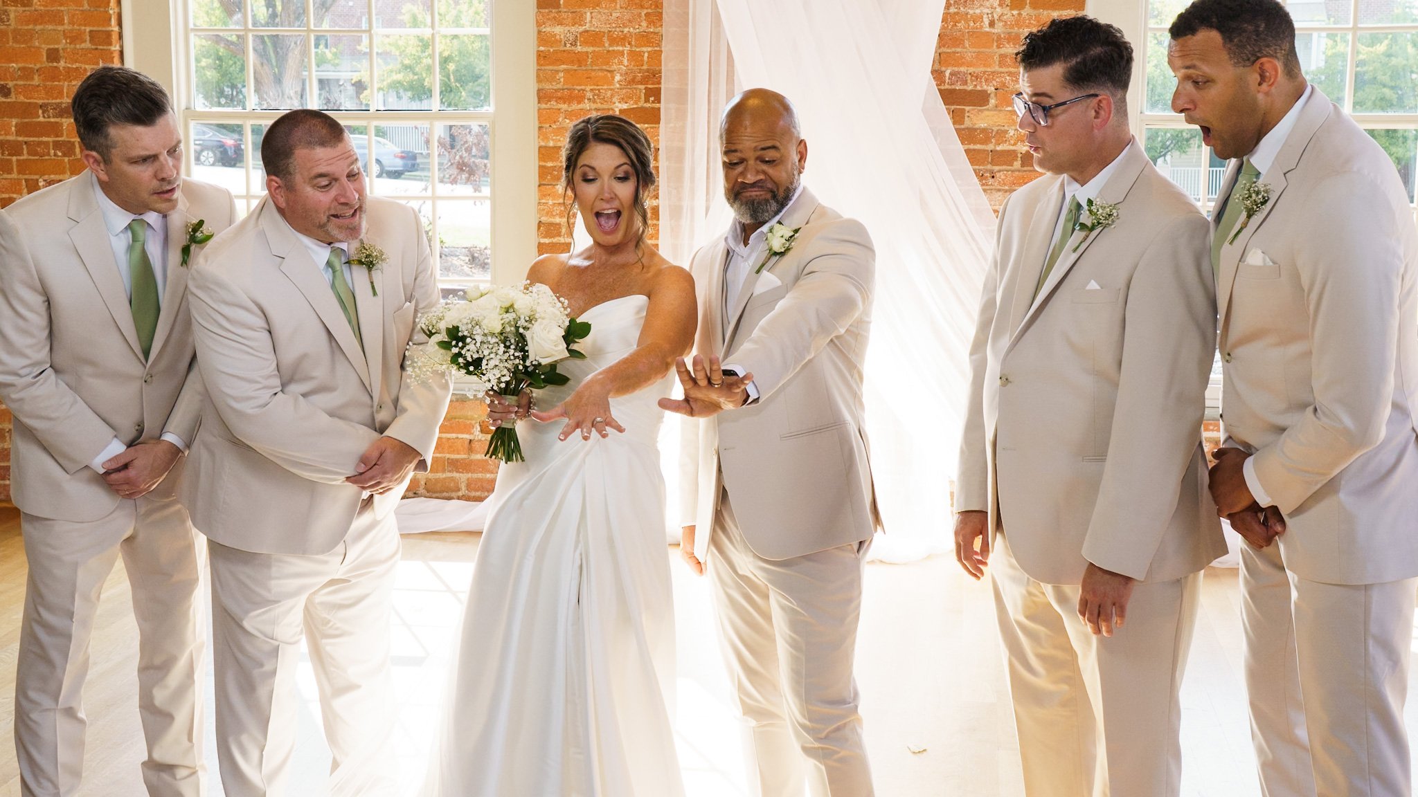Bride showing her wedding ring to five groomsmen in a bright, rustic indoor venue with exposed brick walls and large windows.