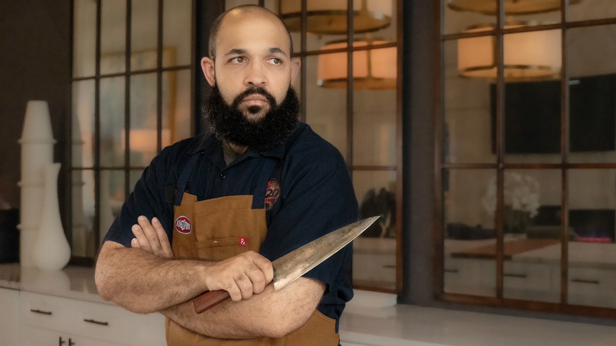 A man with a beard and close-cropped hair, wearing a black shirt and a tan apron, standing in a modern kitchen with arms crossed, holding a metal spatula, looking to the side.