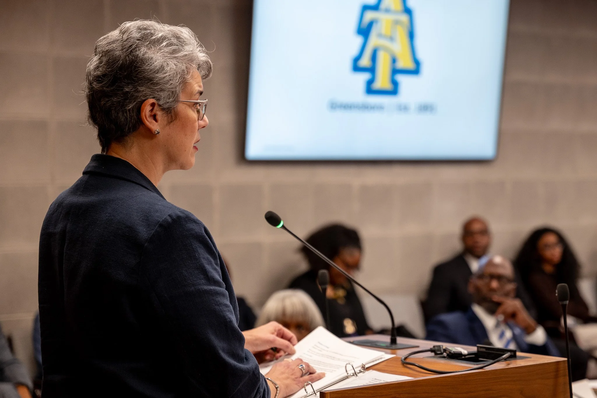 A woman with short gray hair wearing glasses and a dark blazer is speaking at a podium with a microphone. She is holding papers, and there are people seated in the background at an indoor conference or meeting. A large screen behind her displays a lo