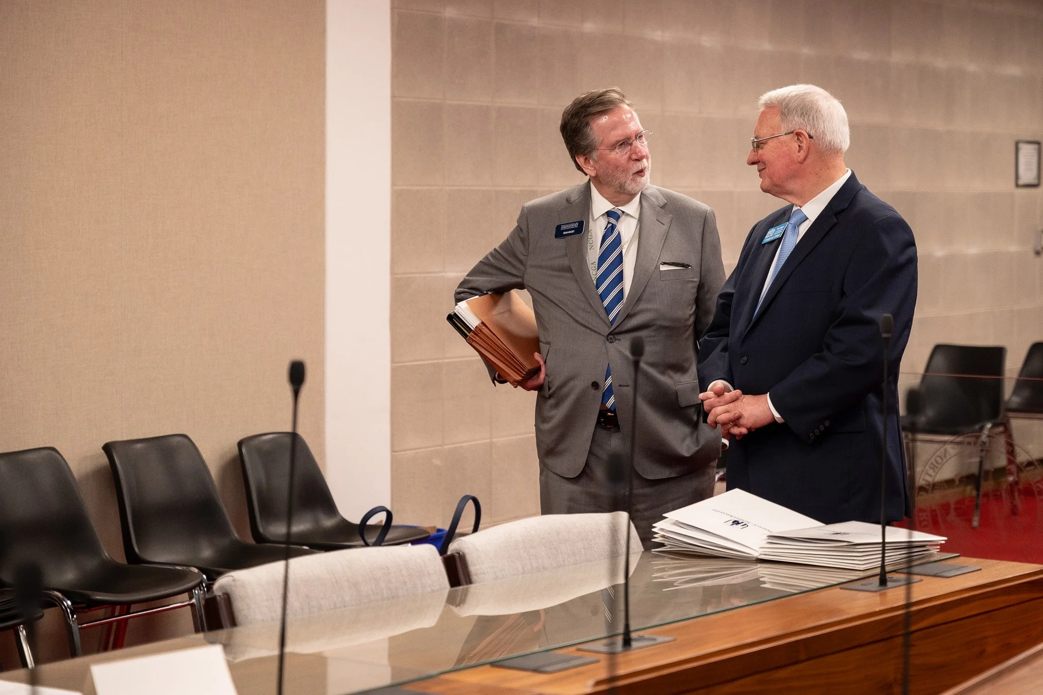 Two men in business suits are having a conversation in a conference room. One is holding folders, and there are chairs, a table with documents, and microphones in the room.