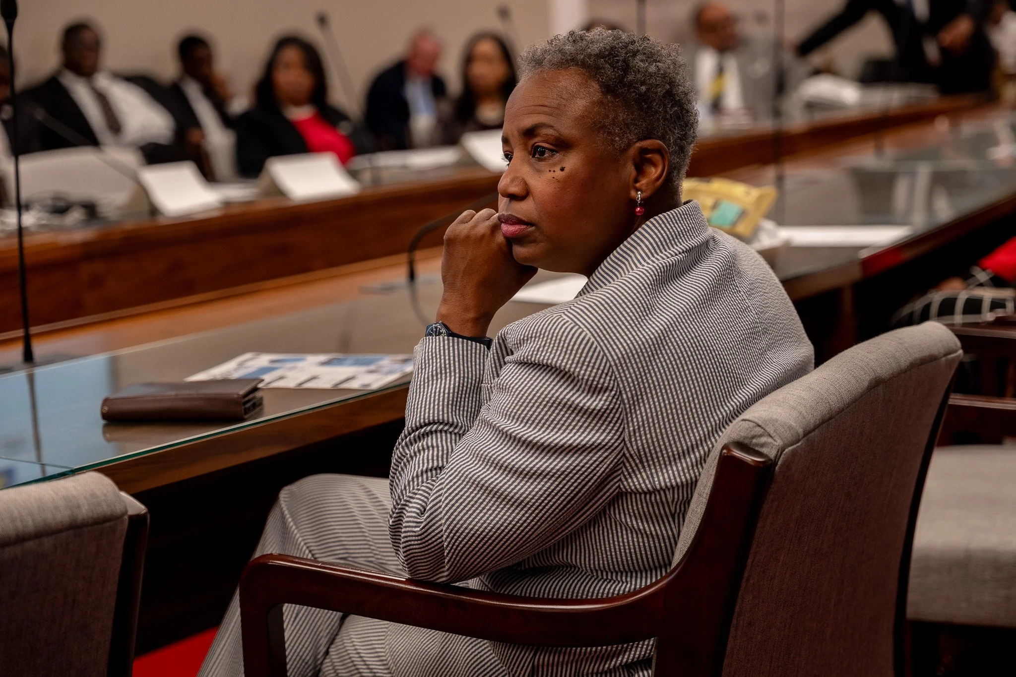 A woman with short gray hair and earrings sitting at a conference table, listening attentively with her chin resting on her hand. Several people are blurred in the background.