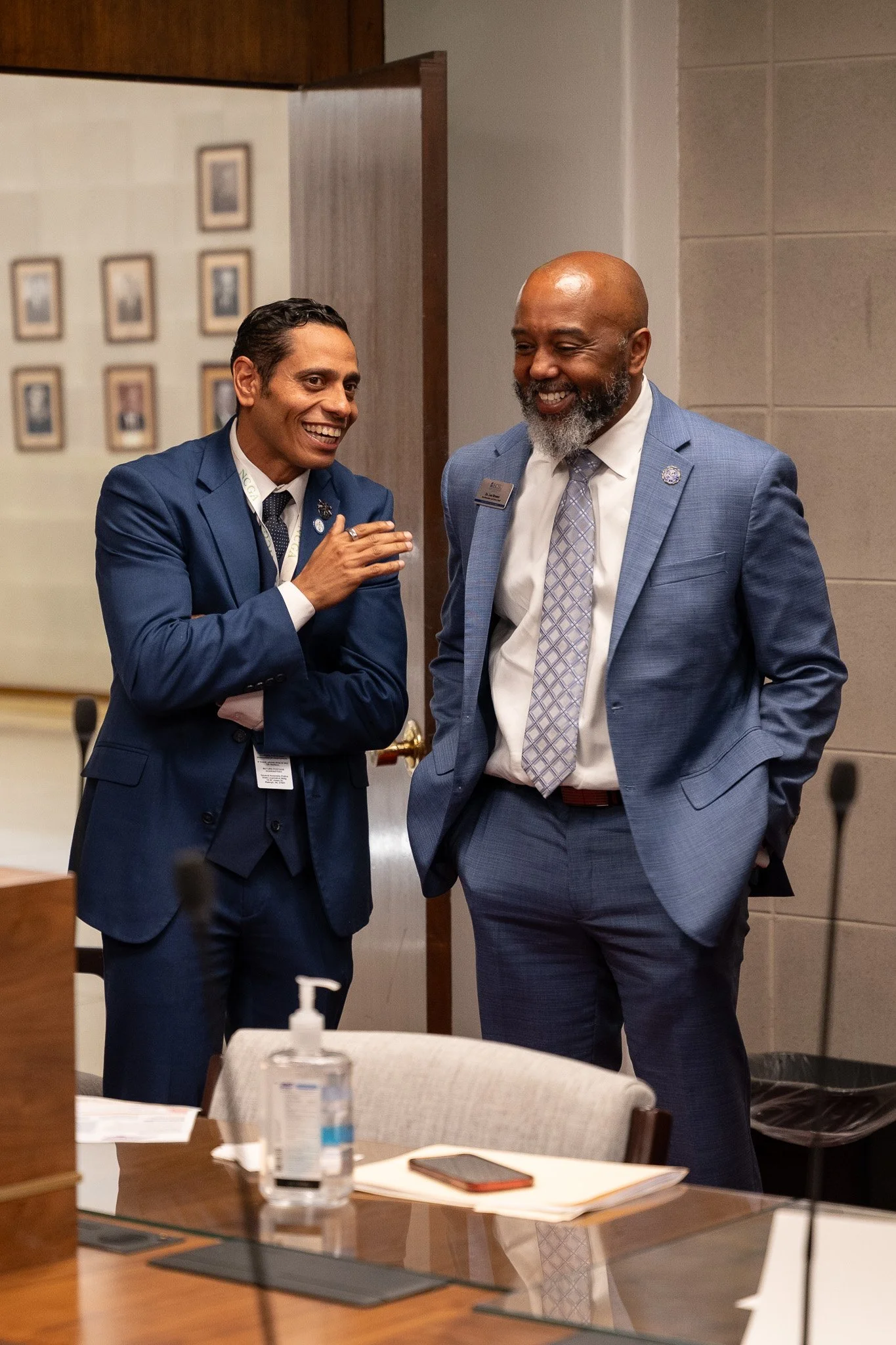 Two men in suits are smiling and talking in a conference room. One has a beard and is bald, the other has dark hair. There are framed pictures on the wall behind them. A table in the foreground has papers, a phone, and a bottle of hand sanitizer.