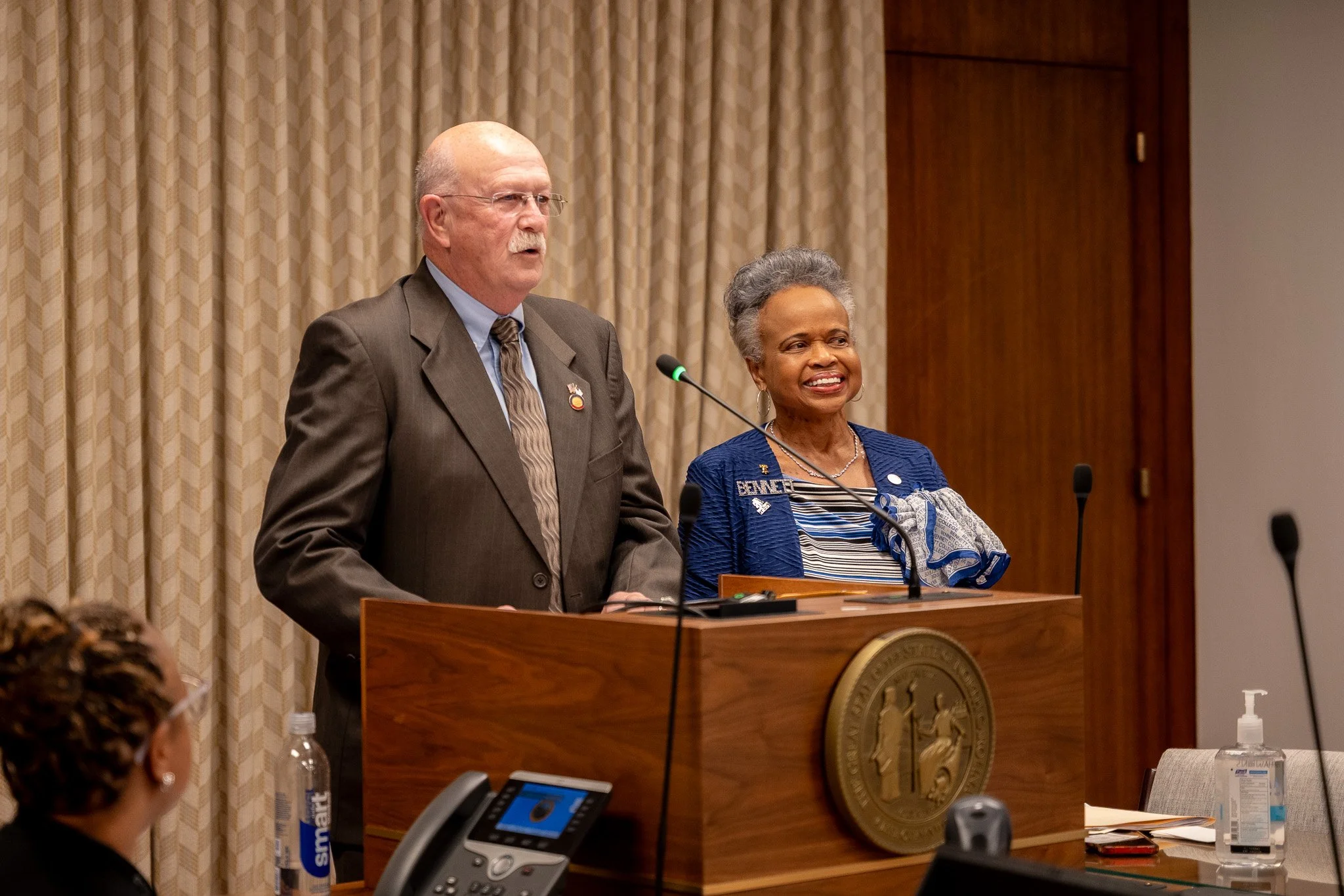 A man and a woman standing at a podium with microphones, giving a speech or presentation in a formal setting.