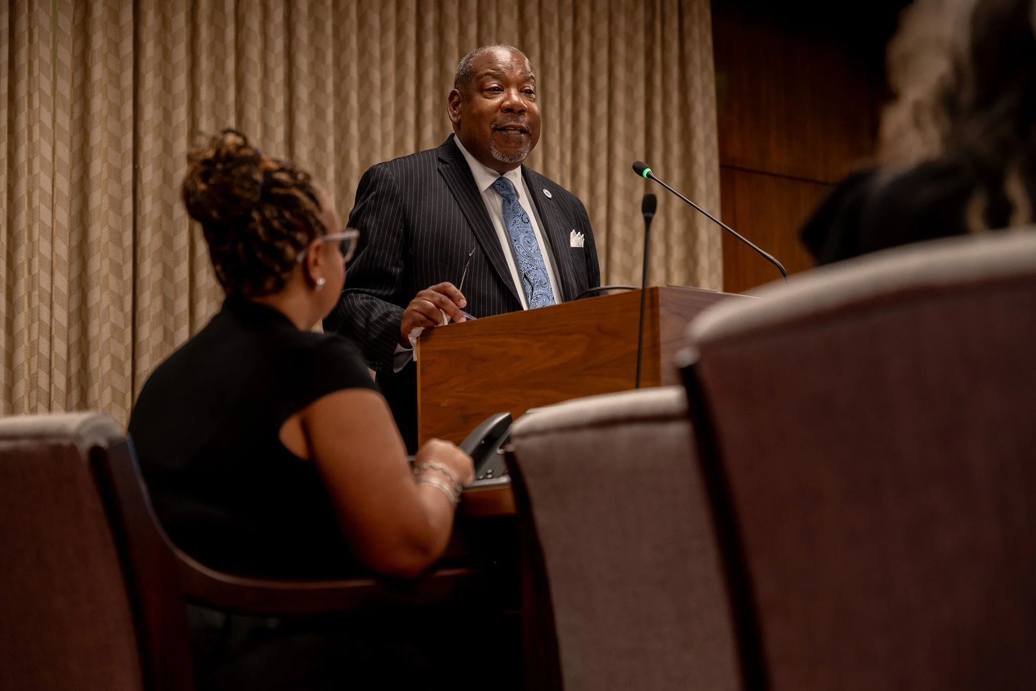 A man in a dark pinstripe suit speaking at a podium during a formal event, with two women seated in front of him listening.
