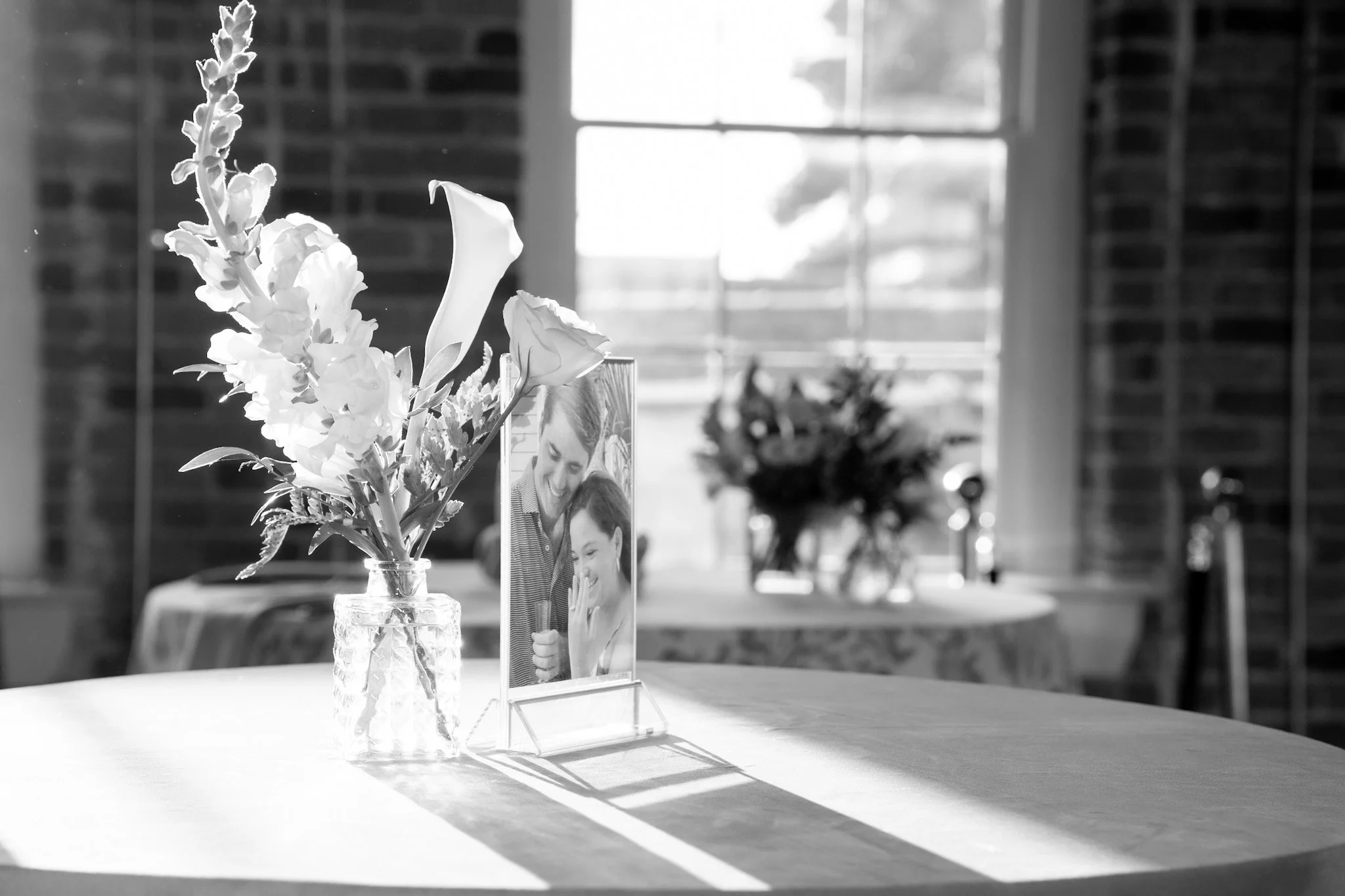 A small glass vase with flowers and a framed photo of a smiling woman and child on a table by a window with sunlight shining through.