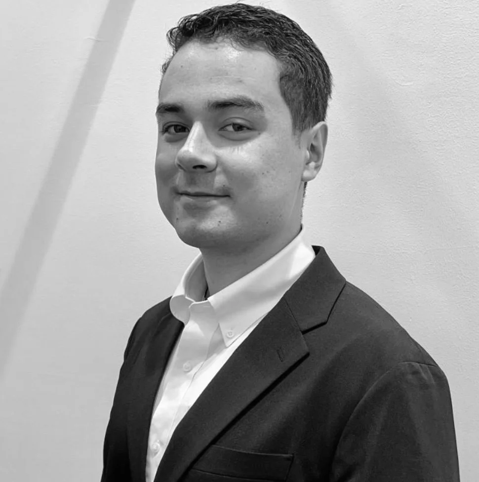 Black and white photo of a young man wearing a suit and white collared shirt, standing against a plain wall, smiling slightly at the camera.