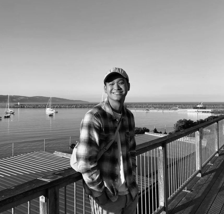 A young man smiling on a wooden pier with boats and a body of water in the background, in black and white.