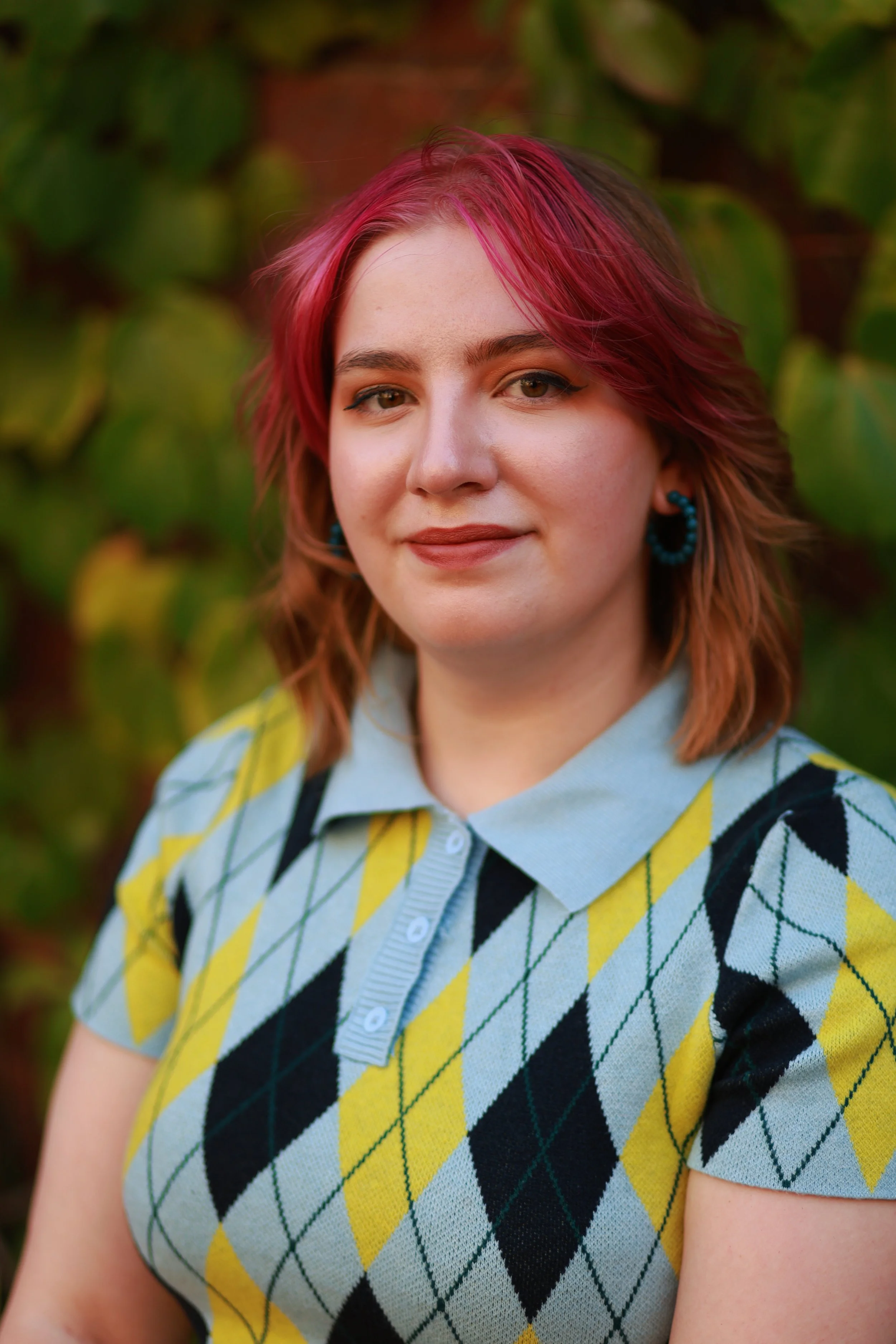 Phoebe, a femme presenting person with pink and auburn hair smiling at the camera in a blue and yellow button up t shirt sweater.