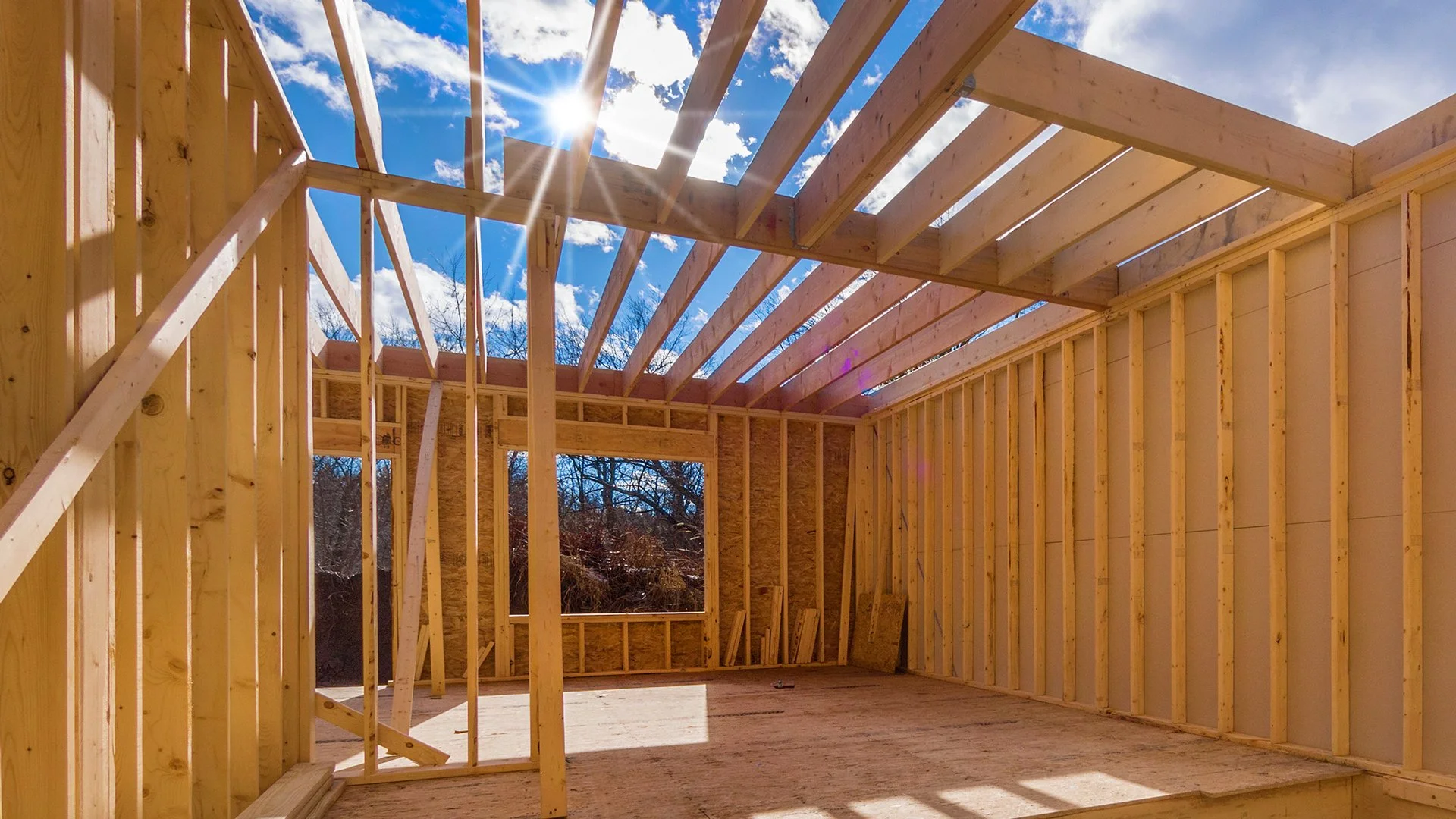 Construction of a home showing the framing and blue sky with sun glaring through the framing