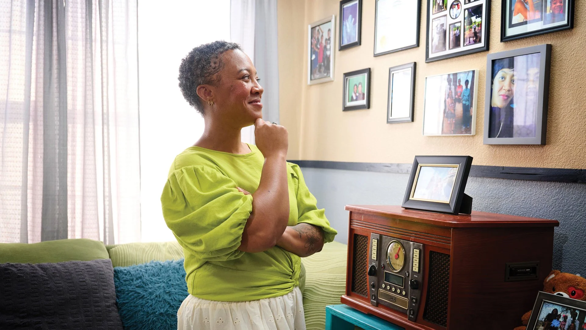 One person standing in a home living room with a pondering look as she looks at a wall of family photos