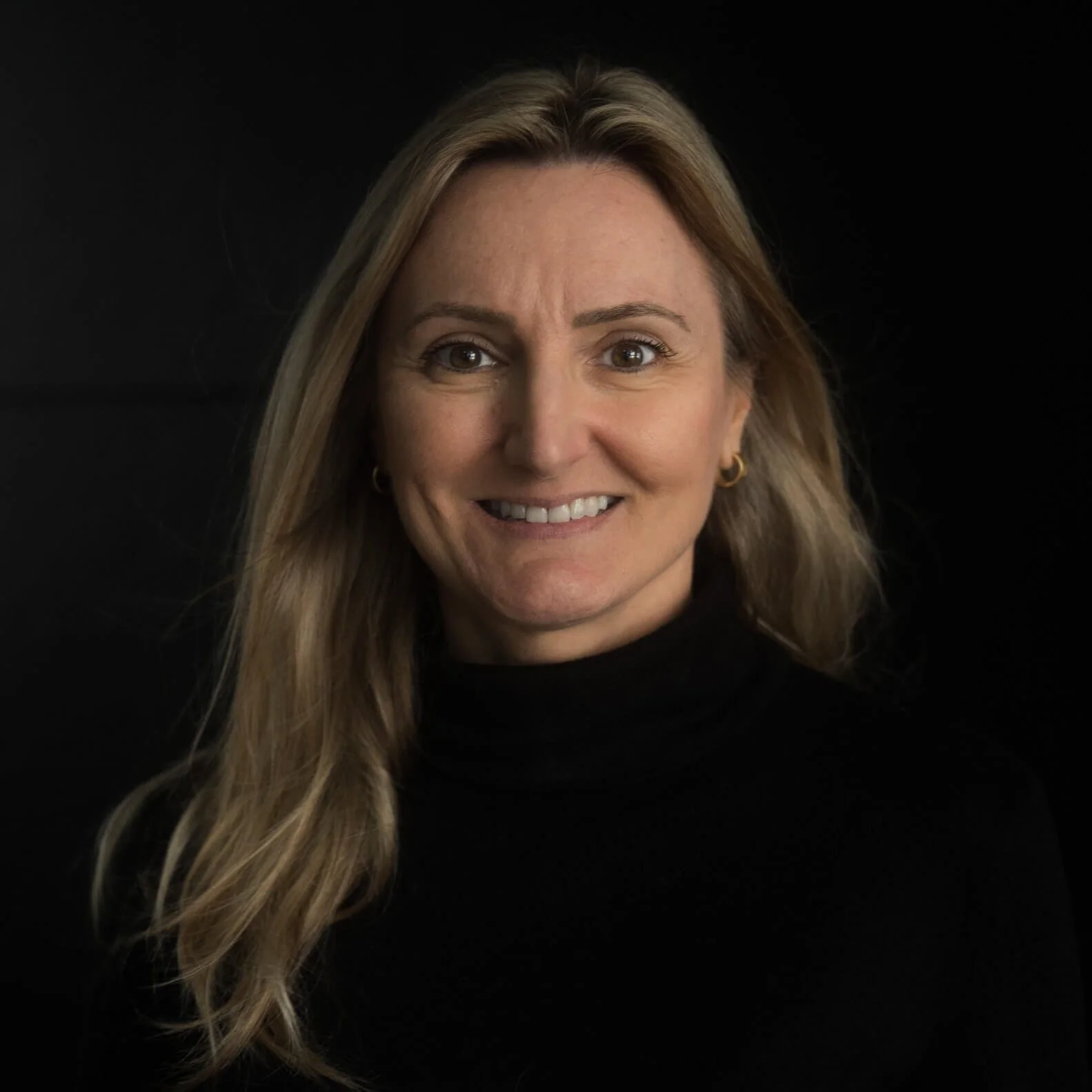 Head and shoulders portrait of a smiling woman with long, wavy blonde hair, wearing a black turtleneck and small gold hoop earrings, against a black background.
