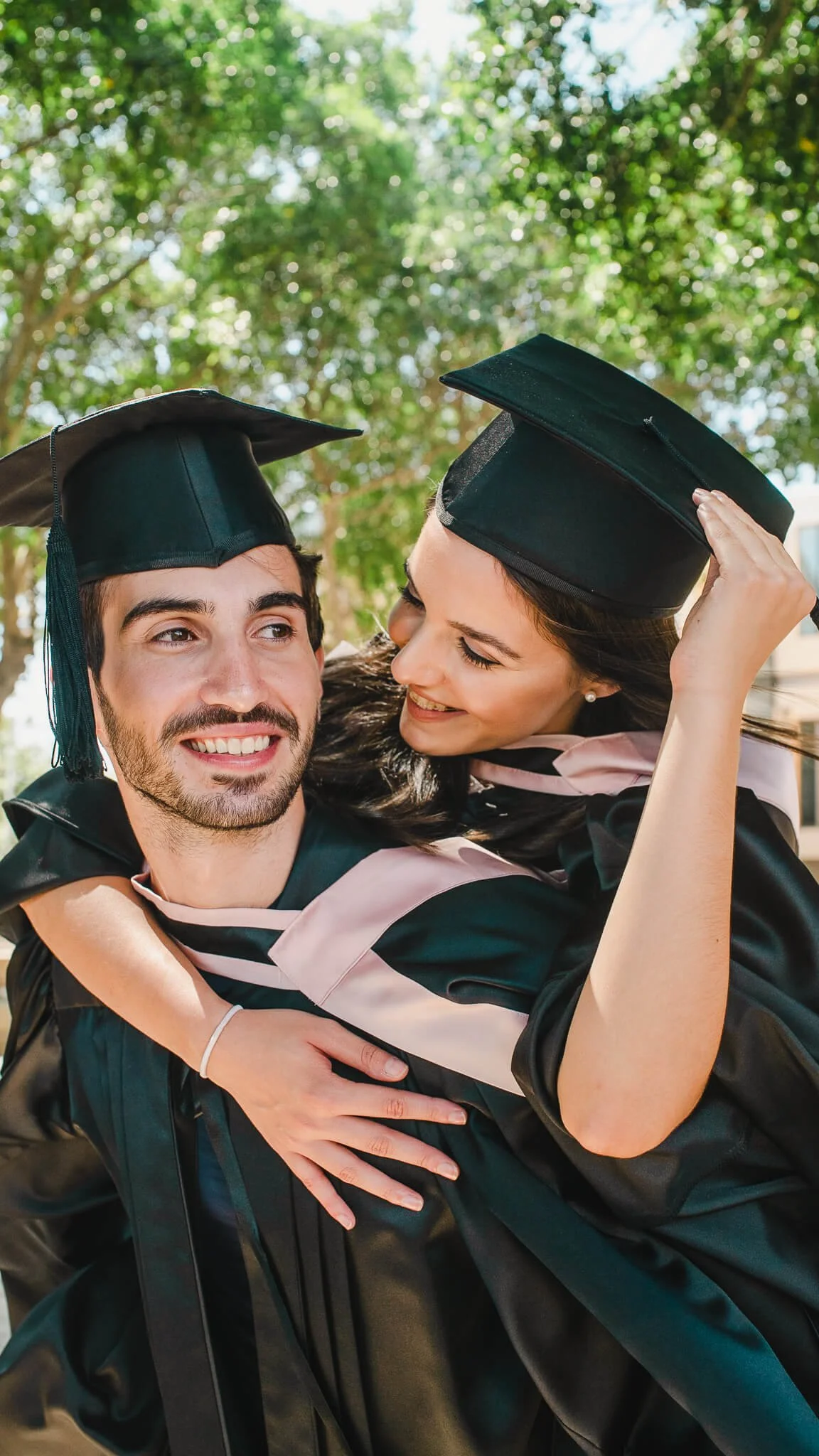 Two graduates in caps and gowns smiling and embracing outdoors on a sunny day.