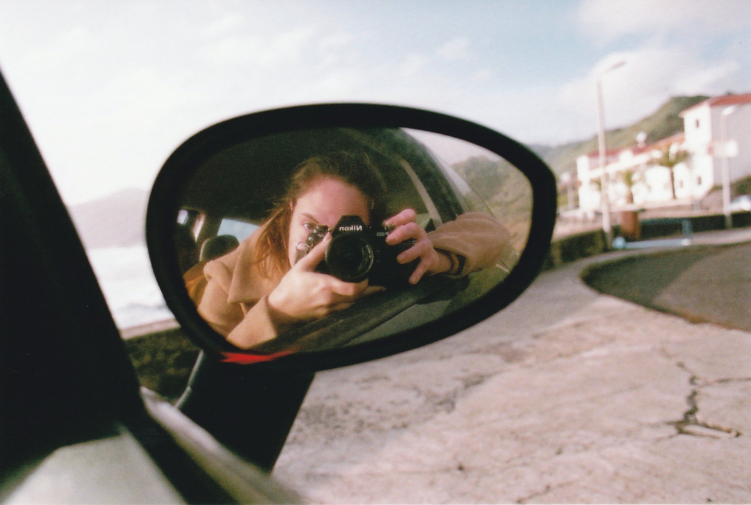 Person taking a photo through a Nikon camera reflected in a car's side mirror, with a road and buildings in the background.