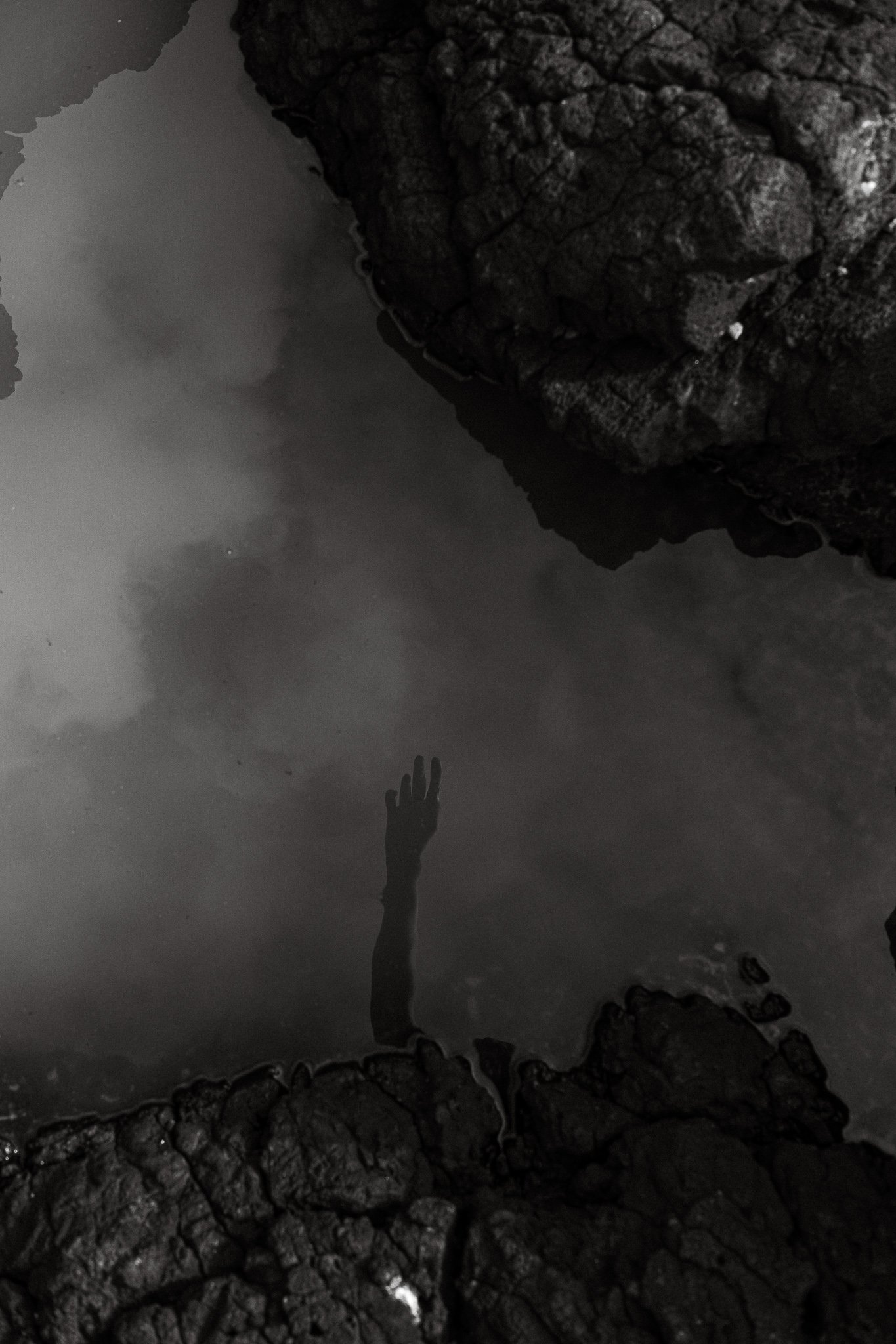 Black and white photo of a hand reaching up through water surrounded by rocks.