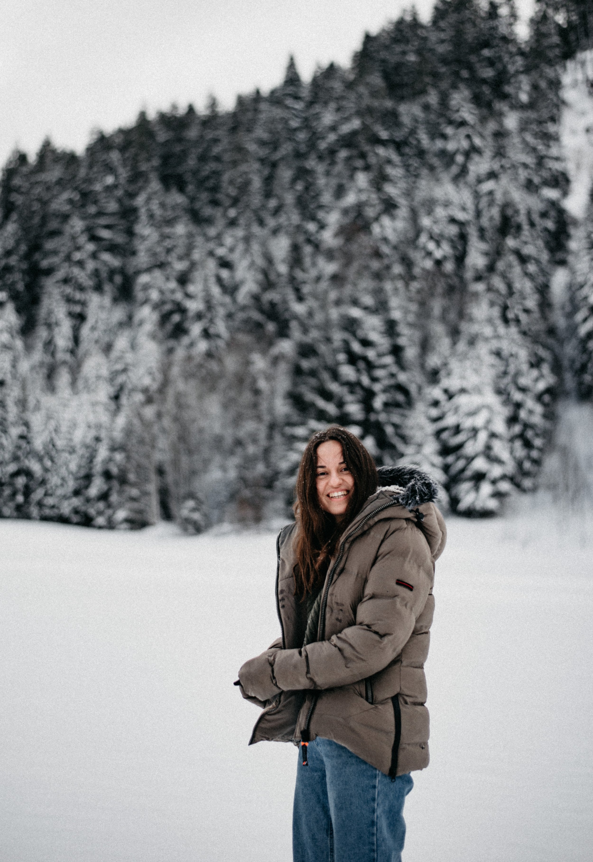 Person wearing a winter jacket standing in a snowy landscape with snow-covered trees in the background.
