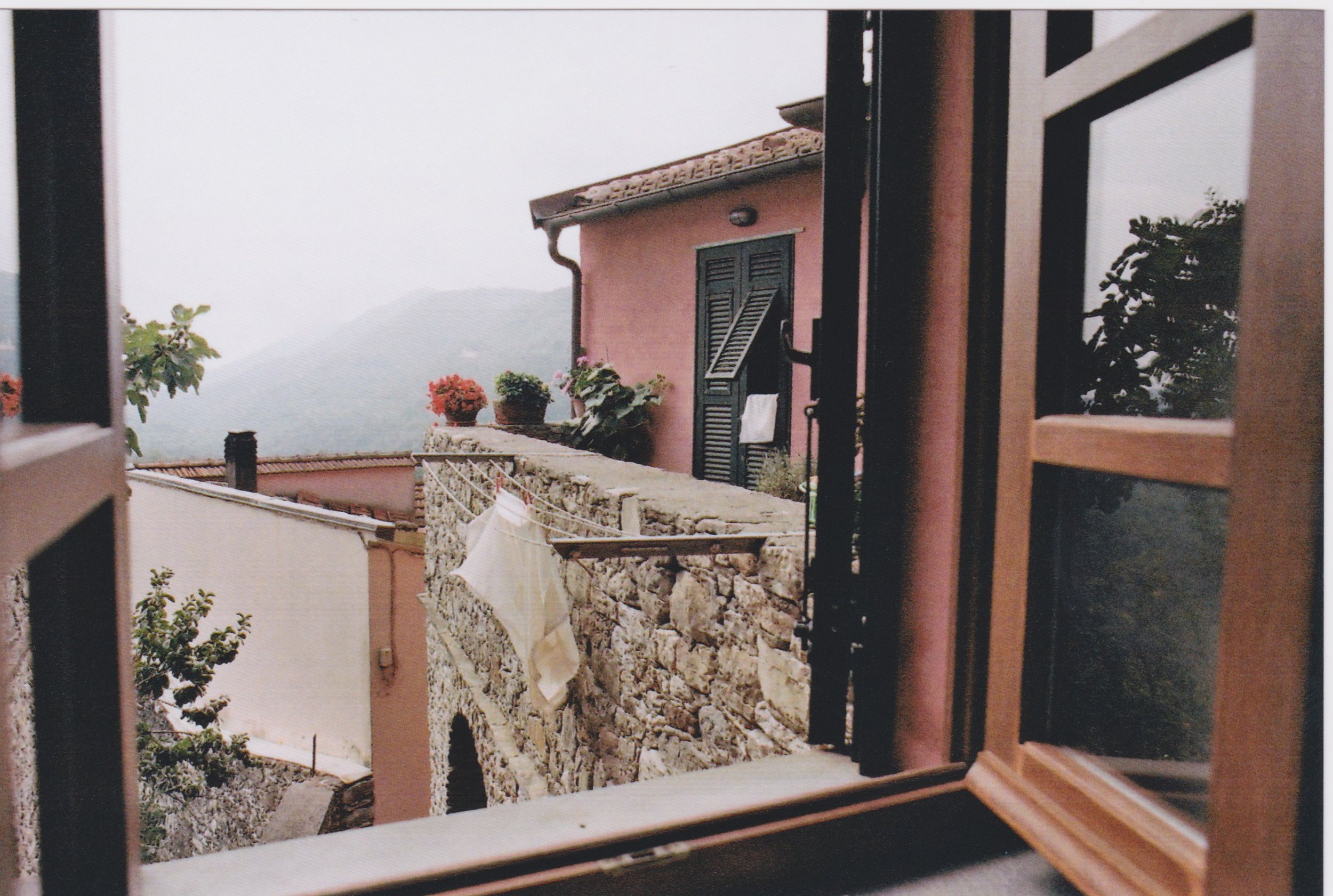 View from a window overlooking a stone balcony with potted plants and drying laundry, scenic hills in the background.
