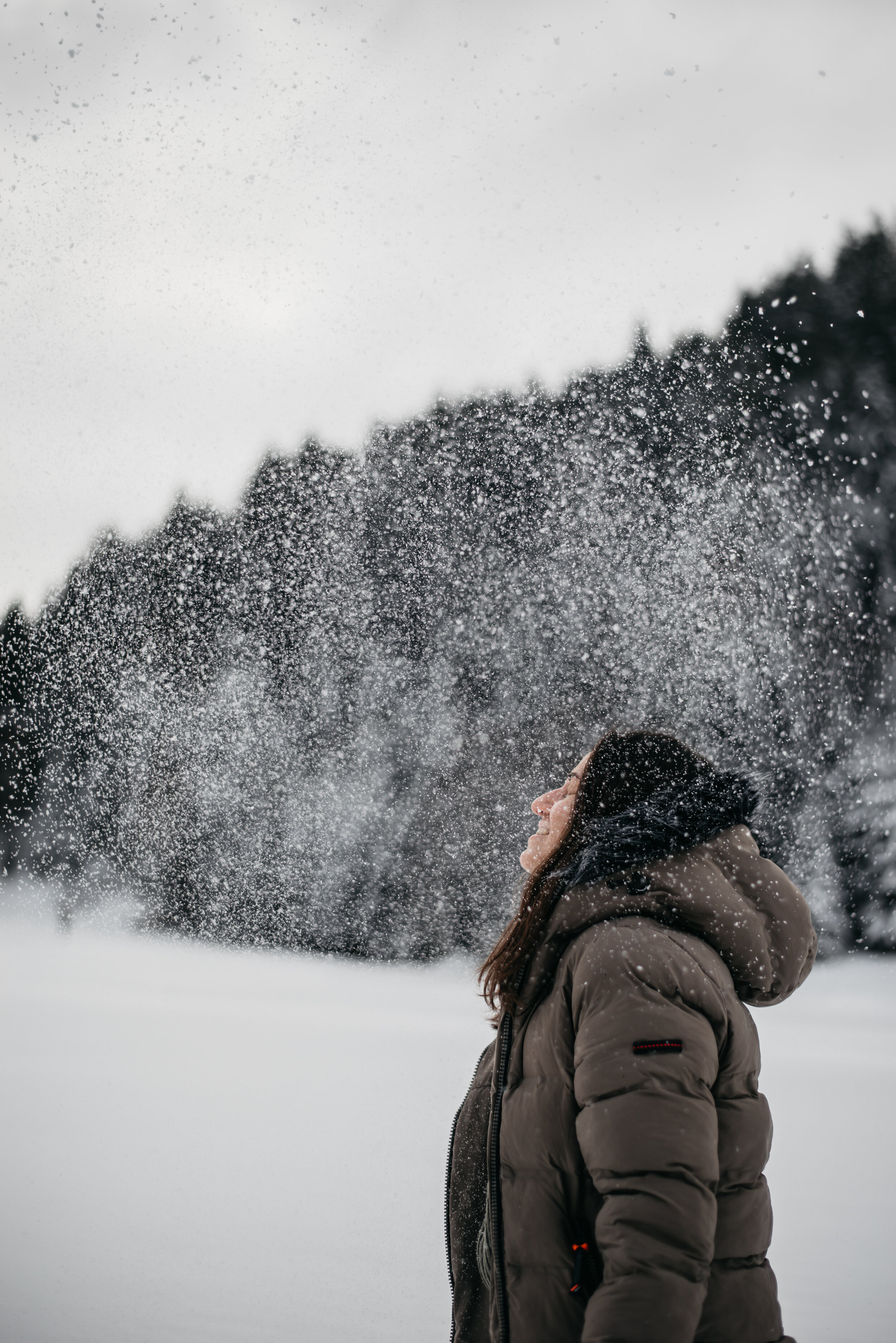 Person in a winter coat standing outdoors in falling snow, surrounded by snow-covered trees.