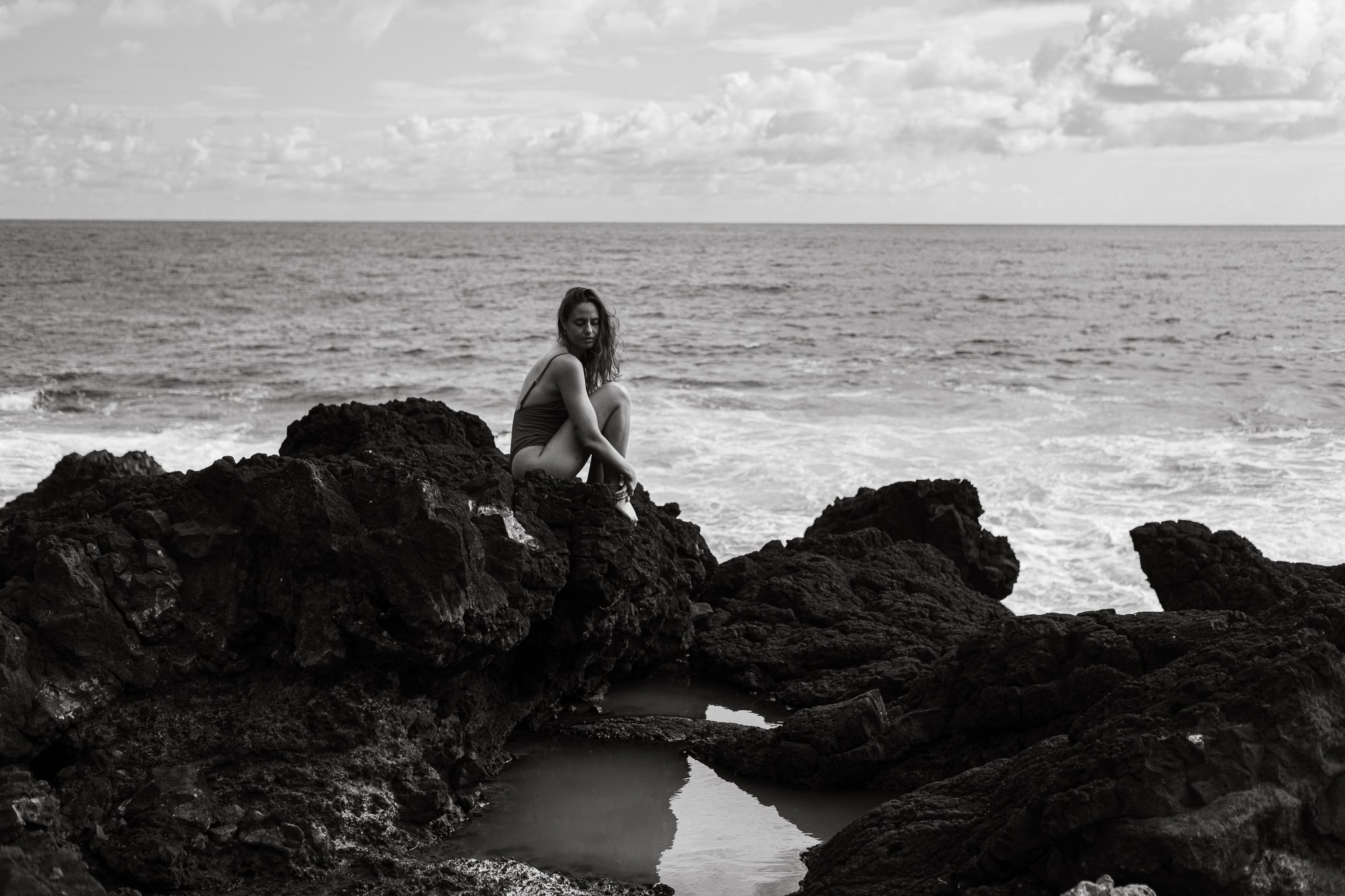 Black and white photo of a person sitting on rocky ocean shore, looking at waves under a cloudy sky.
