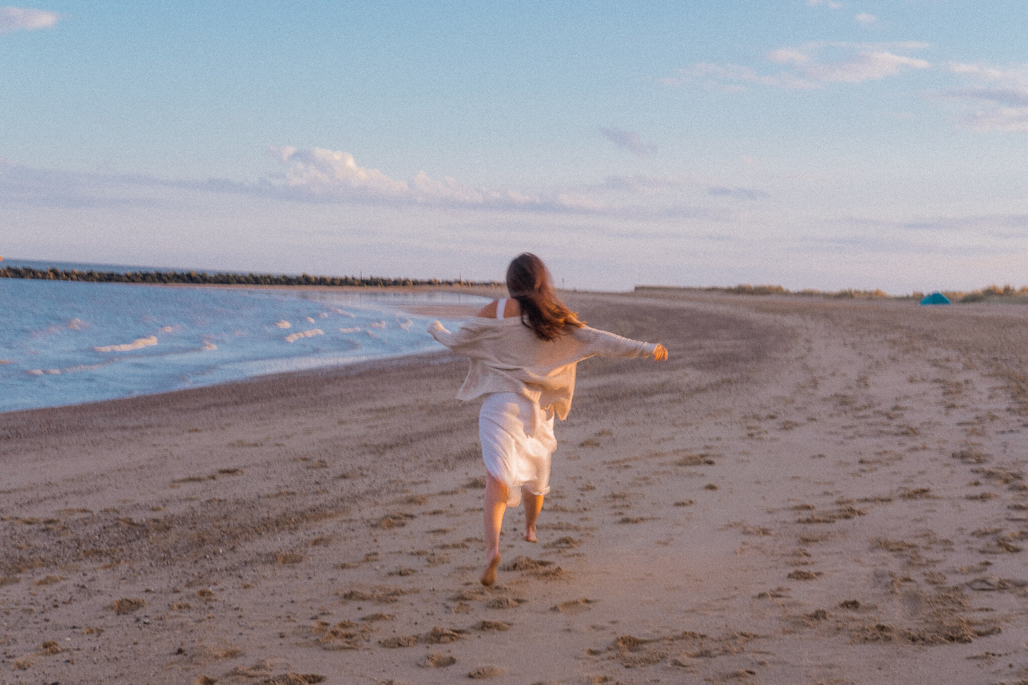 Woman in white dress running along a sandy beach near the ocean.