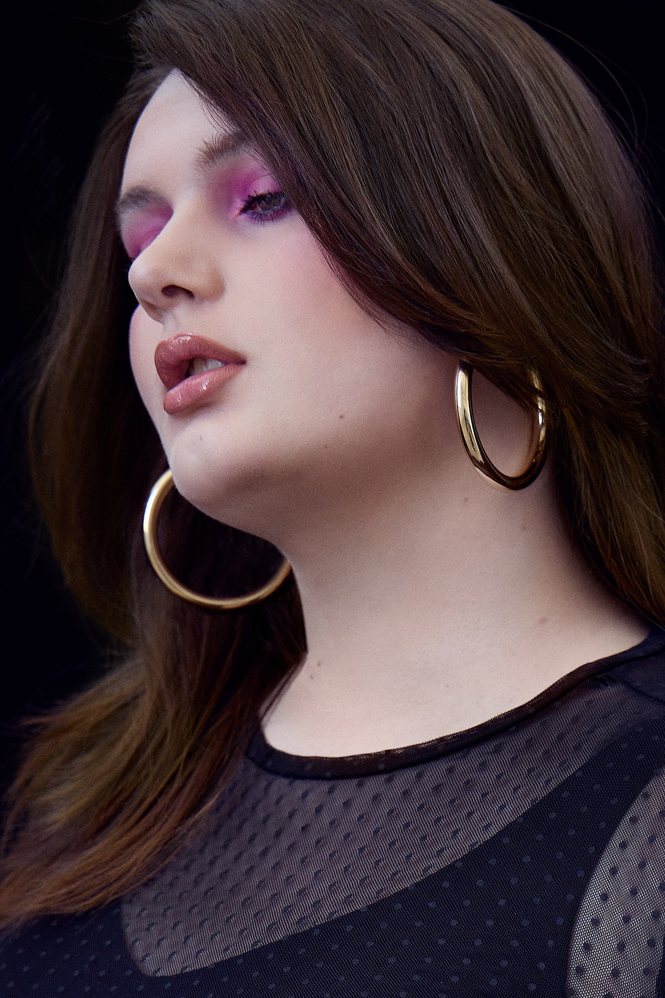 Plus size model with long brown hair, wearing large gold hoop earrings and pink eyeshadow, in a sheer black top.