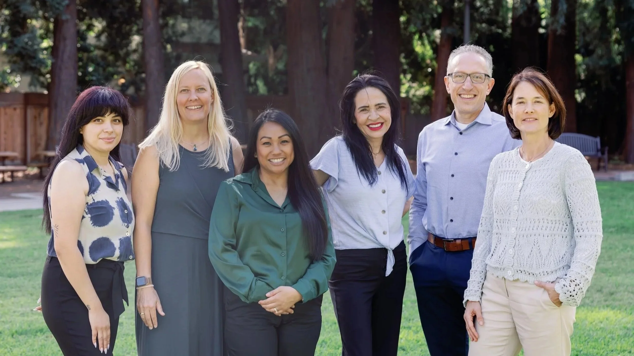 Oakwood Medical team-Group of six diverse adults standing outdoors on grass with trees and a wooden fence in the background.