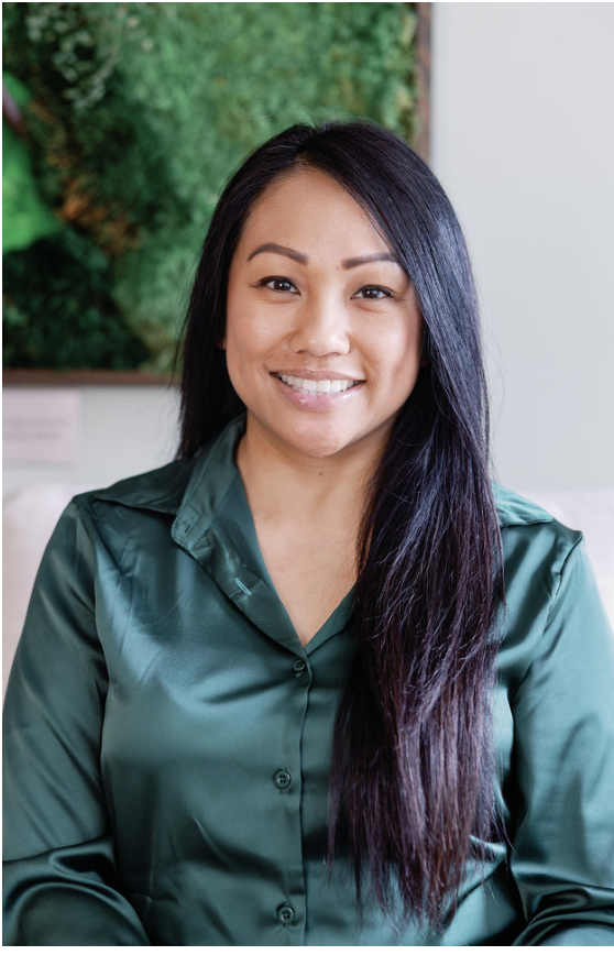 Crystal smiling with long black hair wearing a green satin blouse, sitting indoors with a green plant in the background.