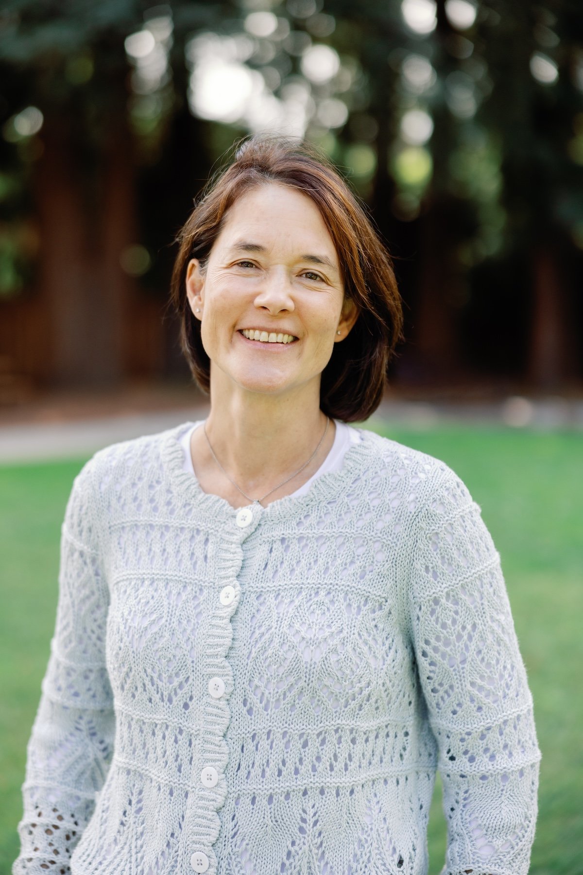Dr. Ando smiling with short brown hair, wearing a light-colored lacy sweater, standing outdoors in a park with trees in the background.