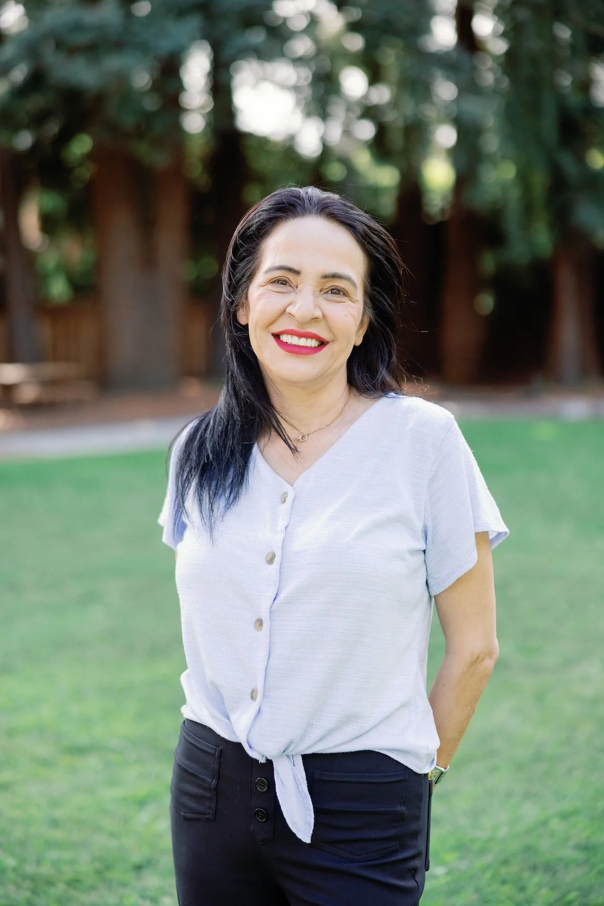 Freba with black hair and red lipstick smiling outdoors on a grassy area, with trees in the background.