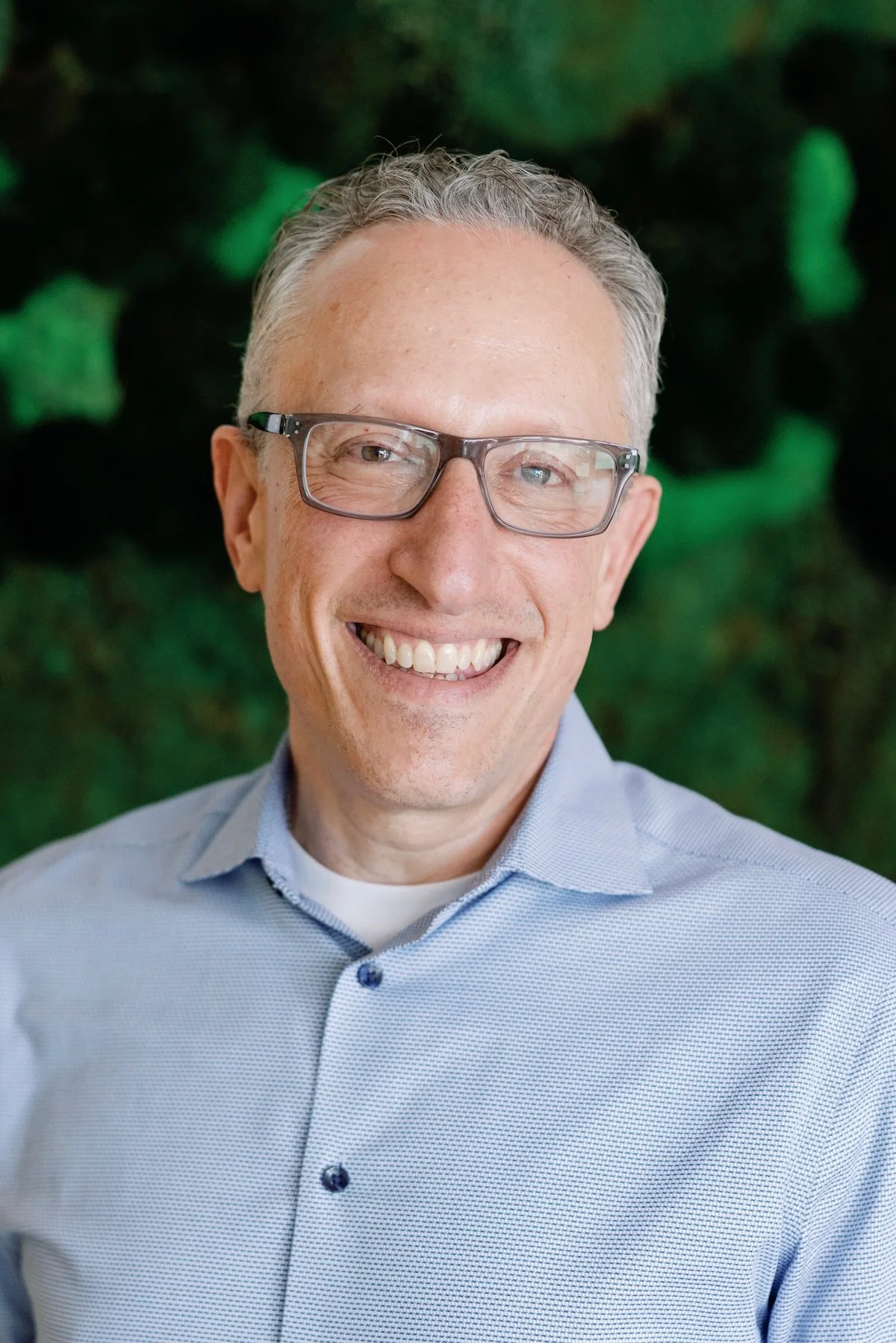 Adam B smiling, with glasses, and a light blue collared shirt standing in front of a green blurred background.