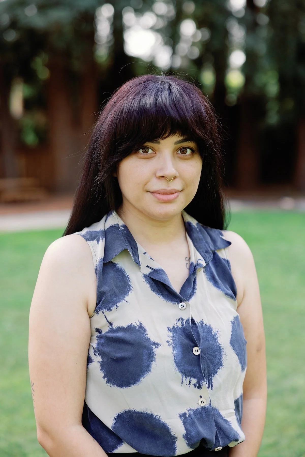 Denise with dark hair, stands outside in a park with grass and trees in the background, wearing a sleeveless beige blouse with dark blue circular patterns.