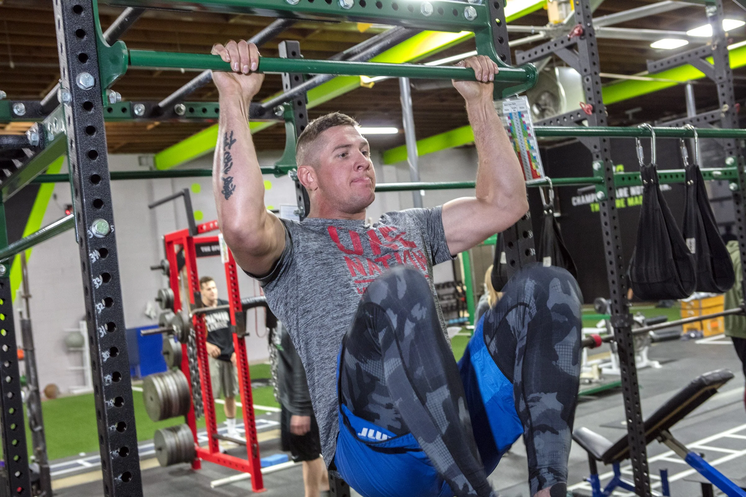 A man doing a pull-up exercise at a gym, surrounded by workout equipment and other individuals in the background.