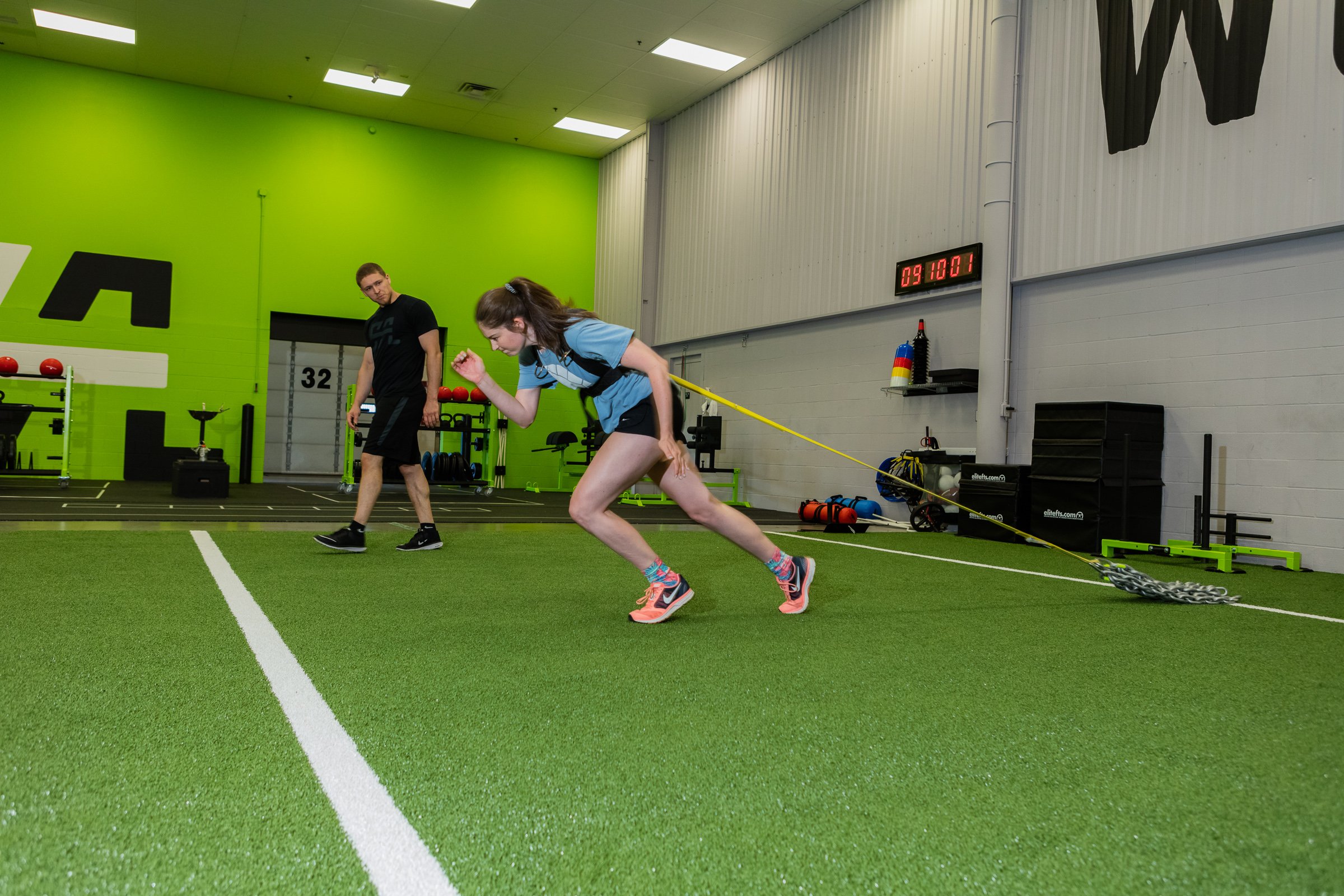 Young woman in athletic clothing pulls resistance bands attached to her waist while training in an indoor gym, with a man observing in the background.