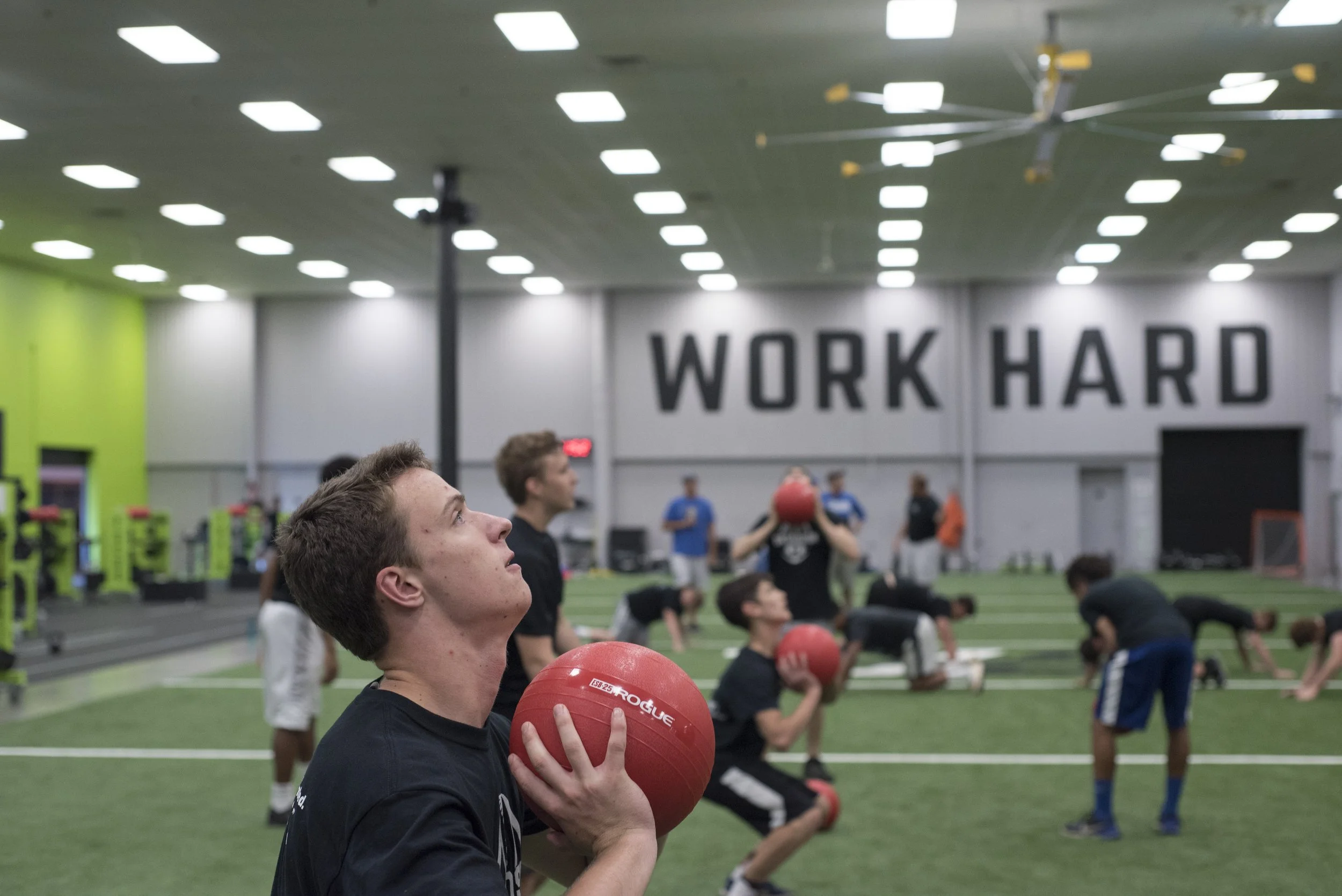 Young athletes in a gym doing medicine ball squats during a workout with a large sign on the wall that says "WORK HARD".