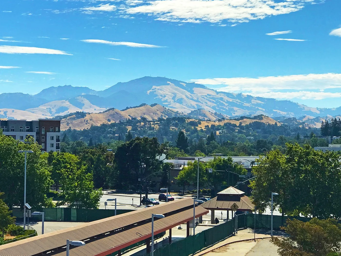 A view of green trees, a parking lot, and a building in the foreground with mountains and blue sky in the background.