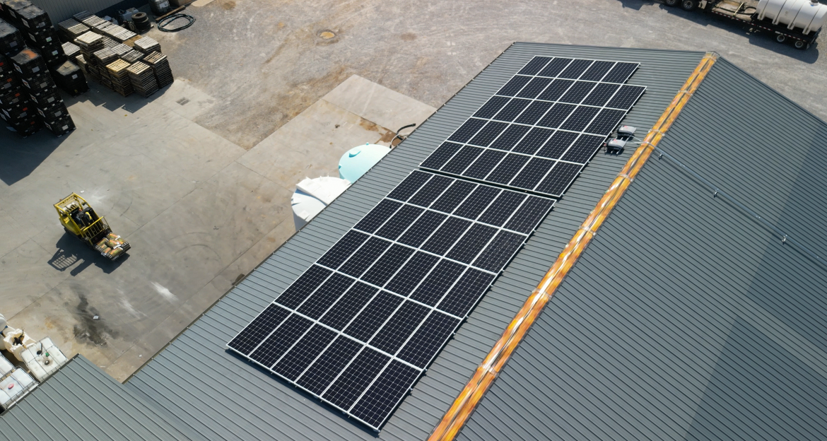Aerial view of a building roof with multiple solar panels installed, and surrounding industrial yard with equipment and storage containers.