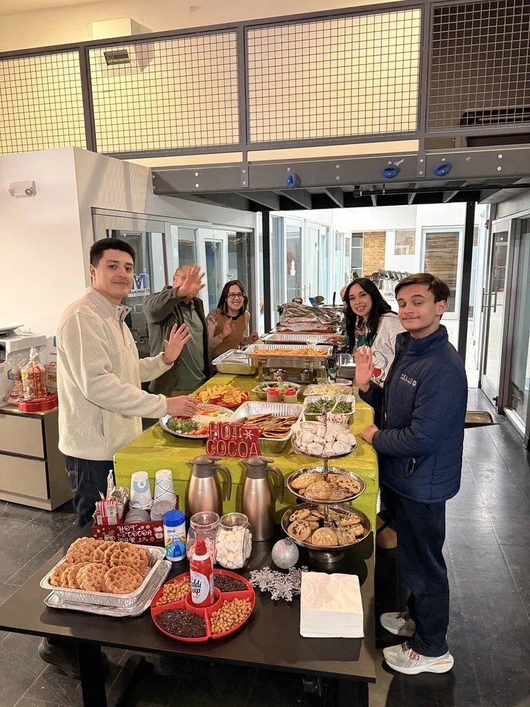 People standing behind a table with various food and desserts, waving at the camera during a holiday gathering.