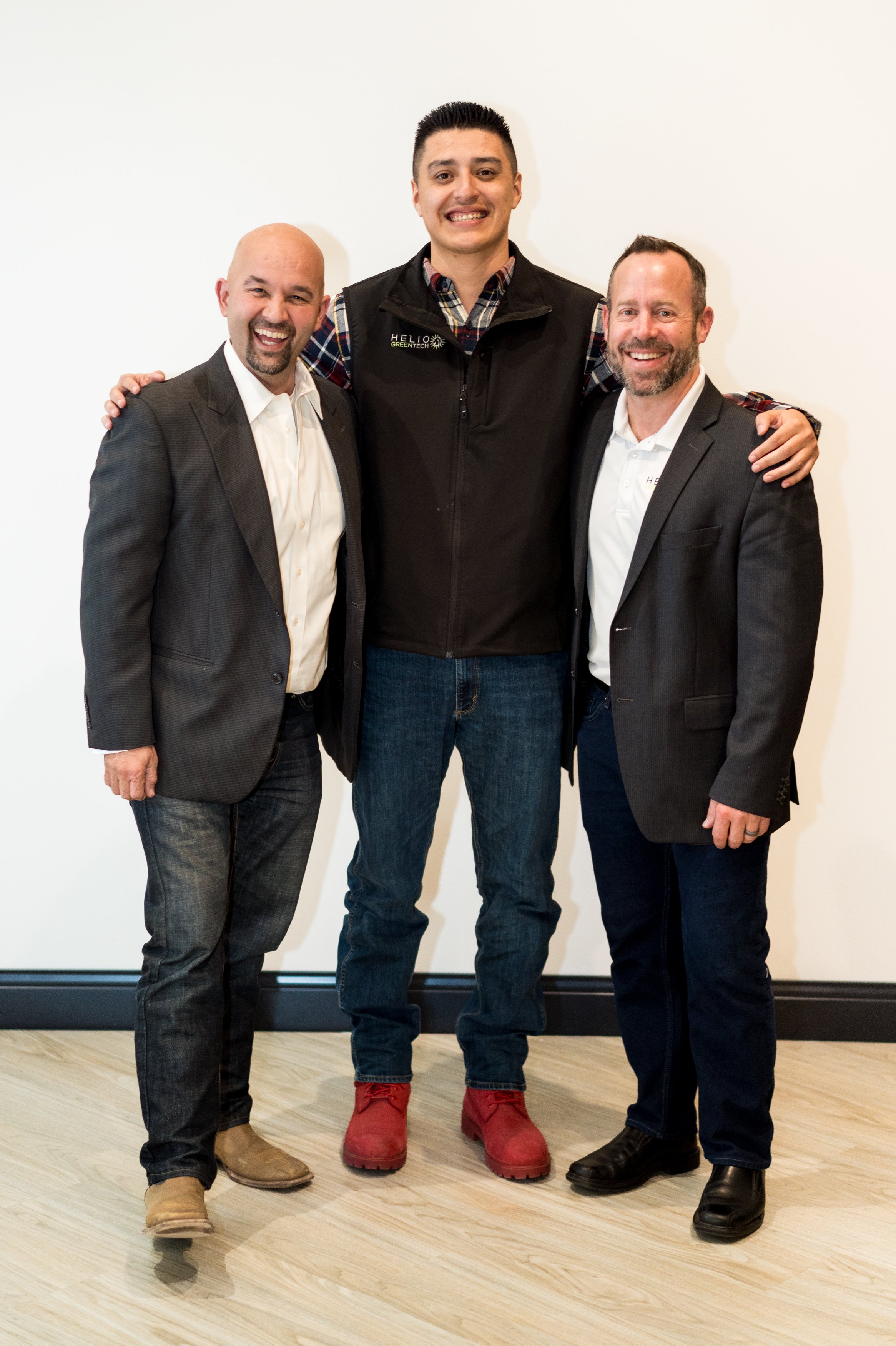 Three men standing together and smiling, with the two men on the sides wearing suits and the man in the middle wearing a black vest, a plaid shirt, and red boots, against a plain white wall.