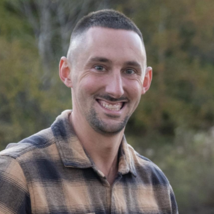 A smiling man with short hair and a beard, wearing a plaid shirt, outdoors with trees in the background.