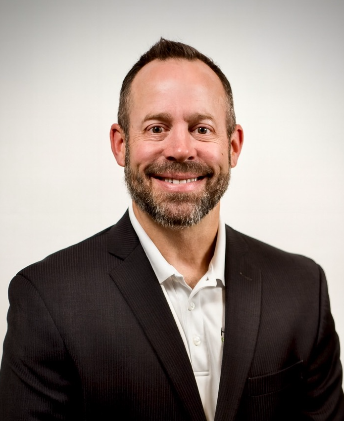 Professional portrait of a smiling man with short dark hair, a beard, and mustache, wearing a black suit jacket and white shirt, against a plain white background.