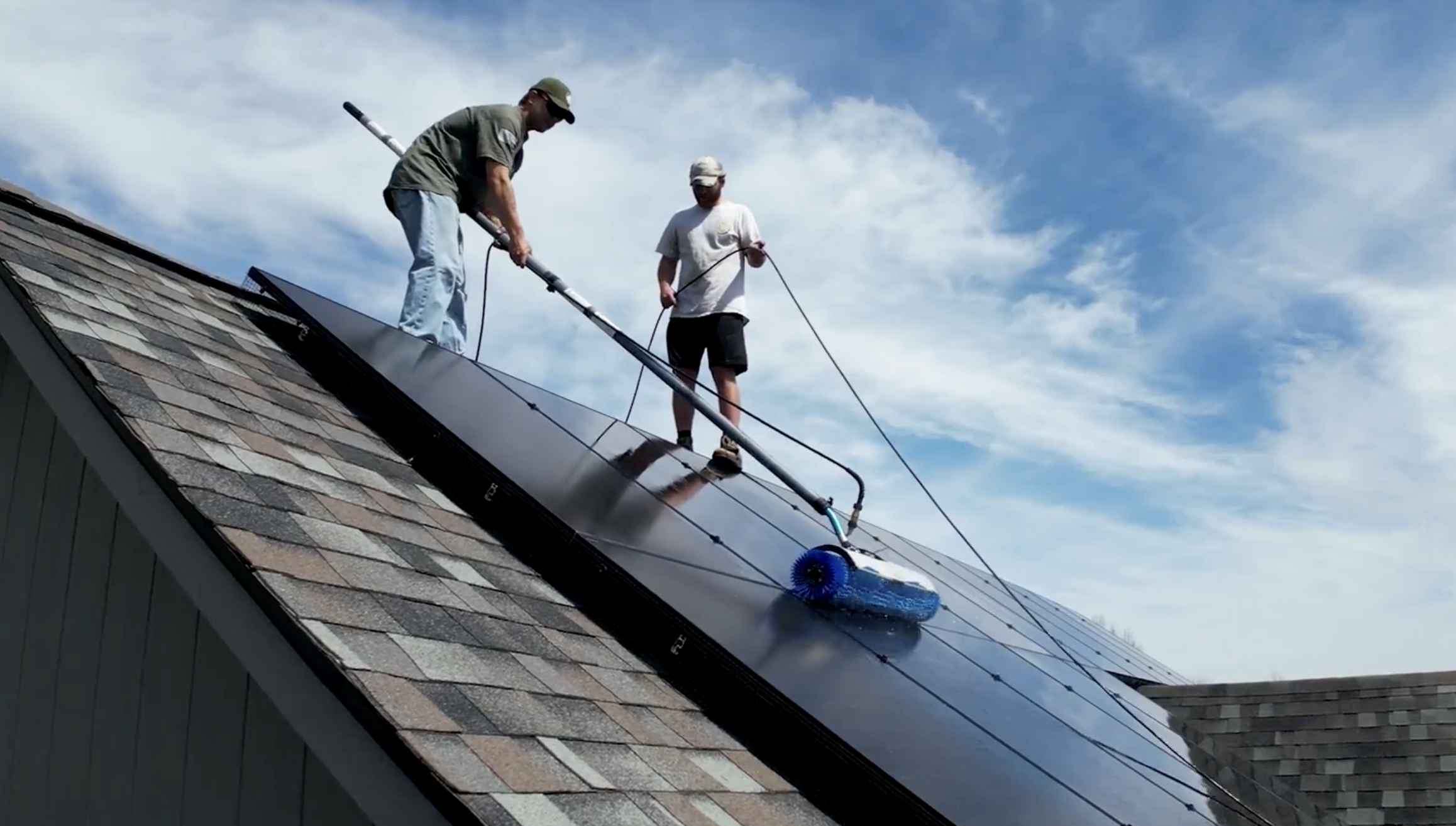 Two men installing or cleaning solar panels on a sloped roof with shingles, using a roller to clean the panels under a blue sky with clouds.