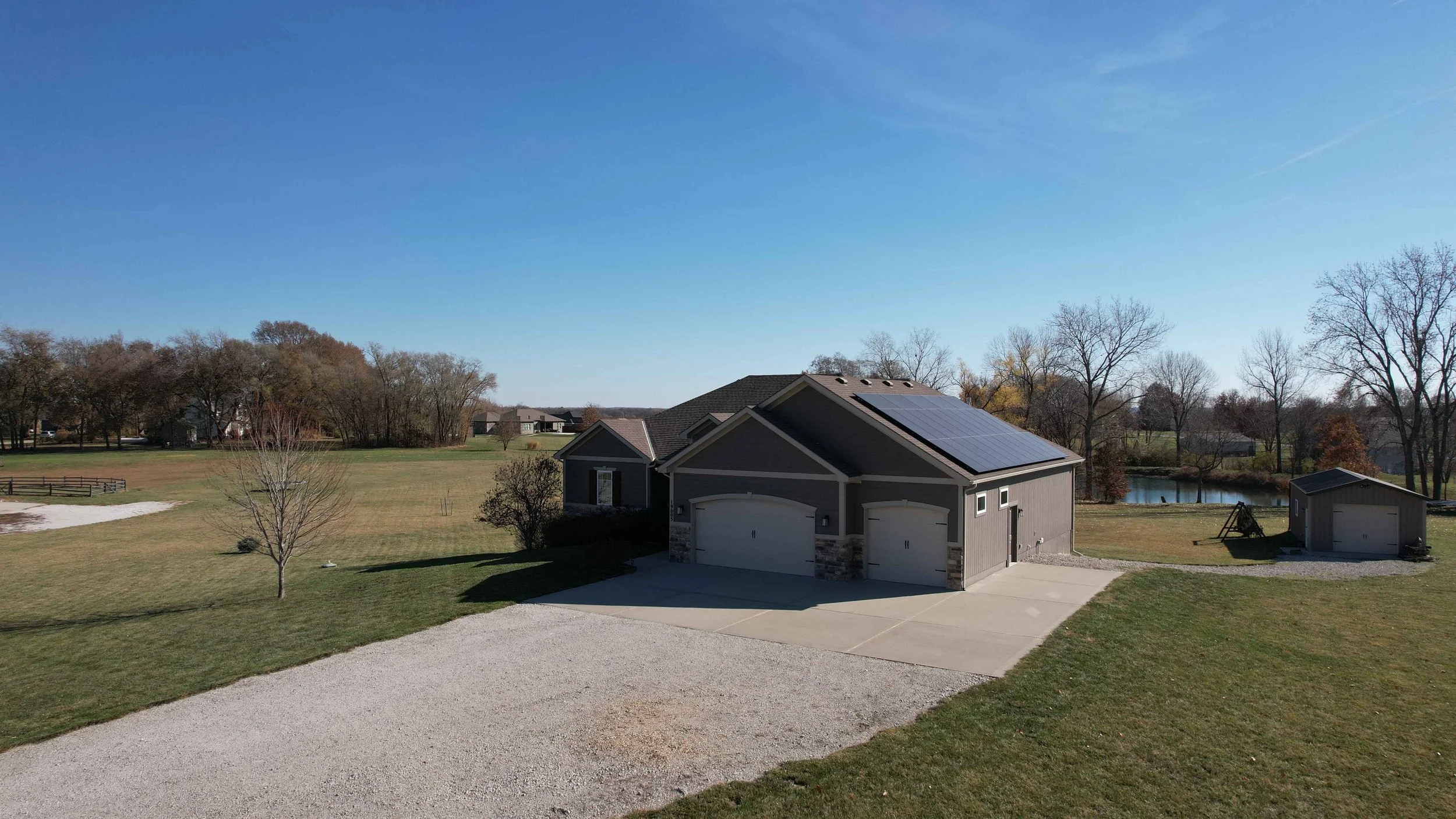 A house with solar panels on the roof, surrounded by a large grassy yard, a small pond, and various trees, under a clear blue sky.
