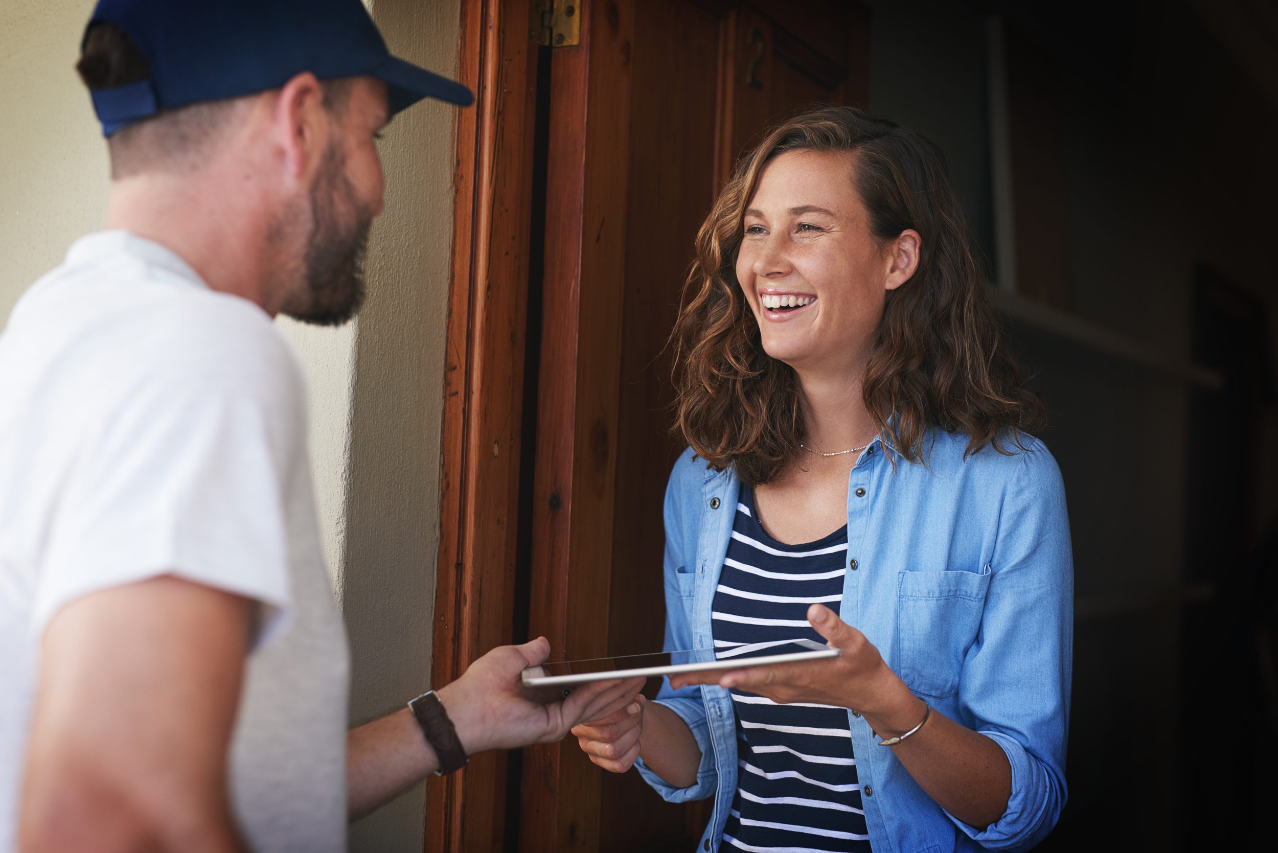 A woman smiling and holding a tablet, standing at a door while a man wearing a cap hands her the device.