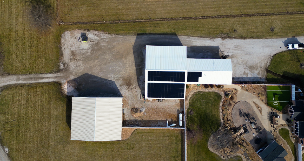 Aerial view of a house with solar panels, a detached garage, a driveway, a small garden, and playground area with a small soccer field.