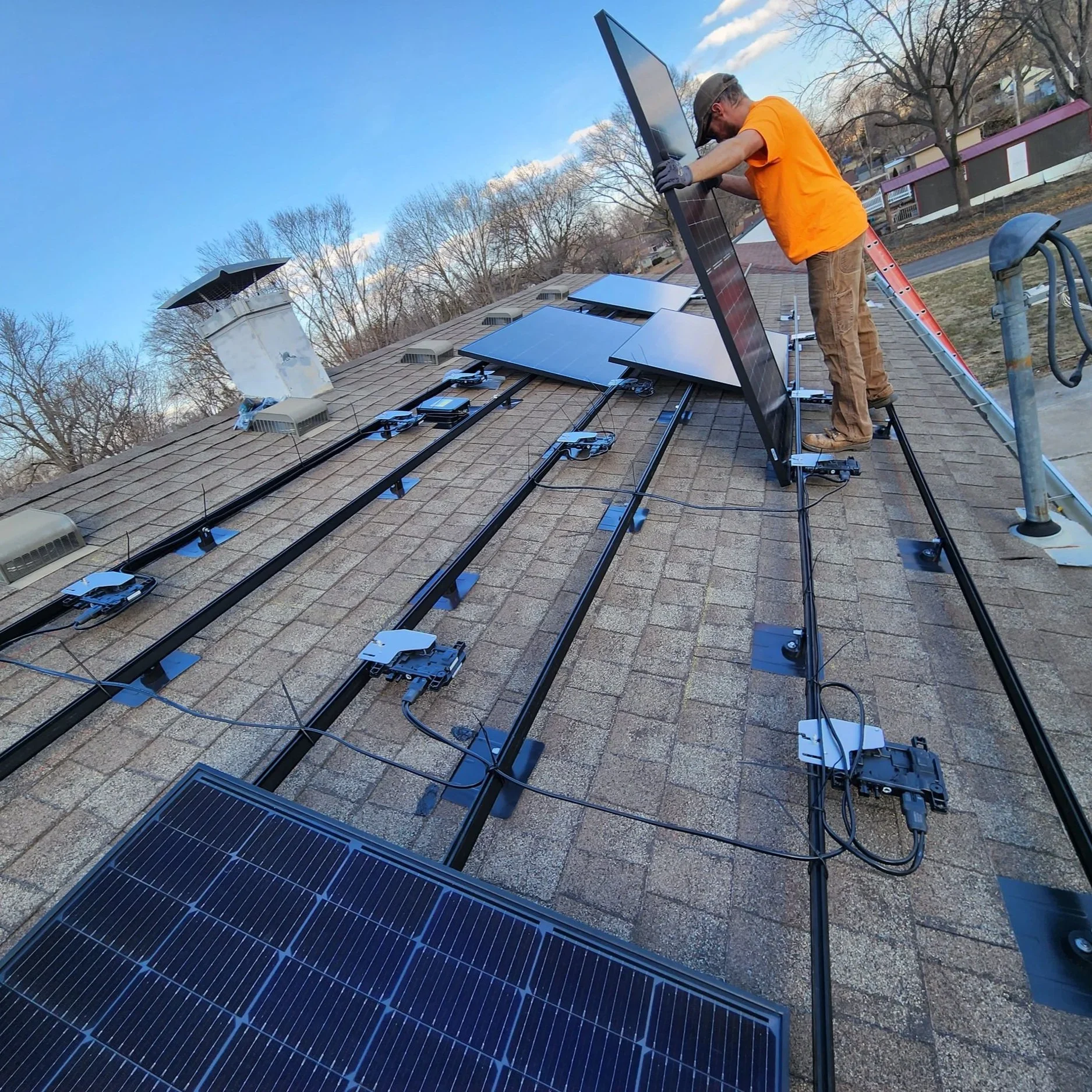 A worker installing solar panels on a house roof during daytime, with tools and mounting hardware visible.