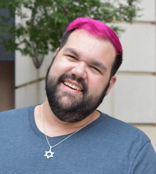 A smiling man with a beard, short dark hair with a pink streak, wearing a blue shirt and a star-shaped necklace, outdoors with trees and a building in the background.